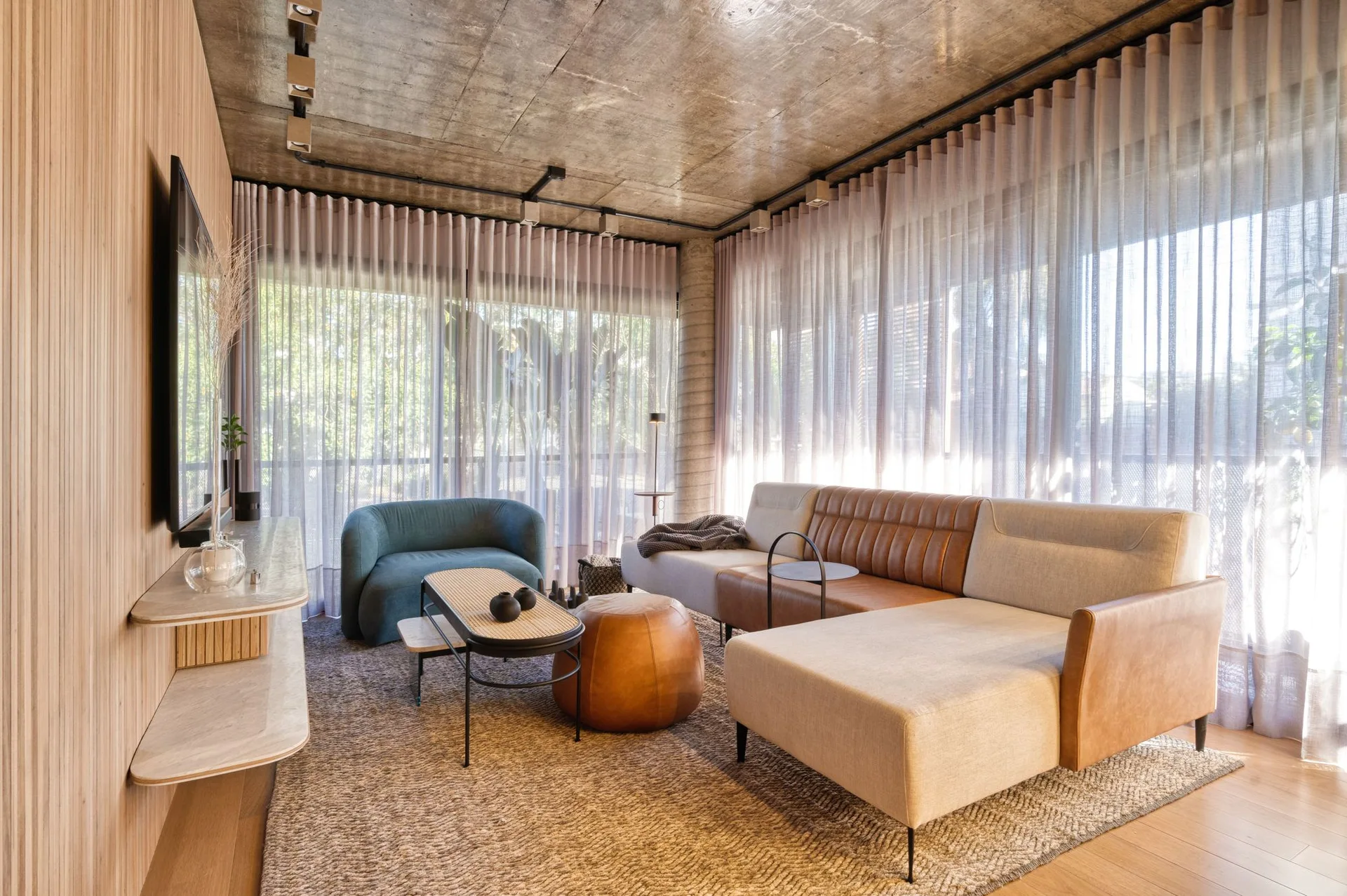 A modern living room featuring a concrete ceiling, vertical wood slat wall, sheer curtains, and a mixed leather and fabric sectional sofa.