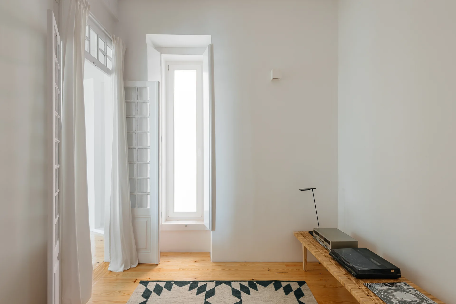 A bright white minimalist room featuring a long woven rattan bench, a geometric patterned rug, and a tall narrow window over light wood floors.