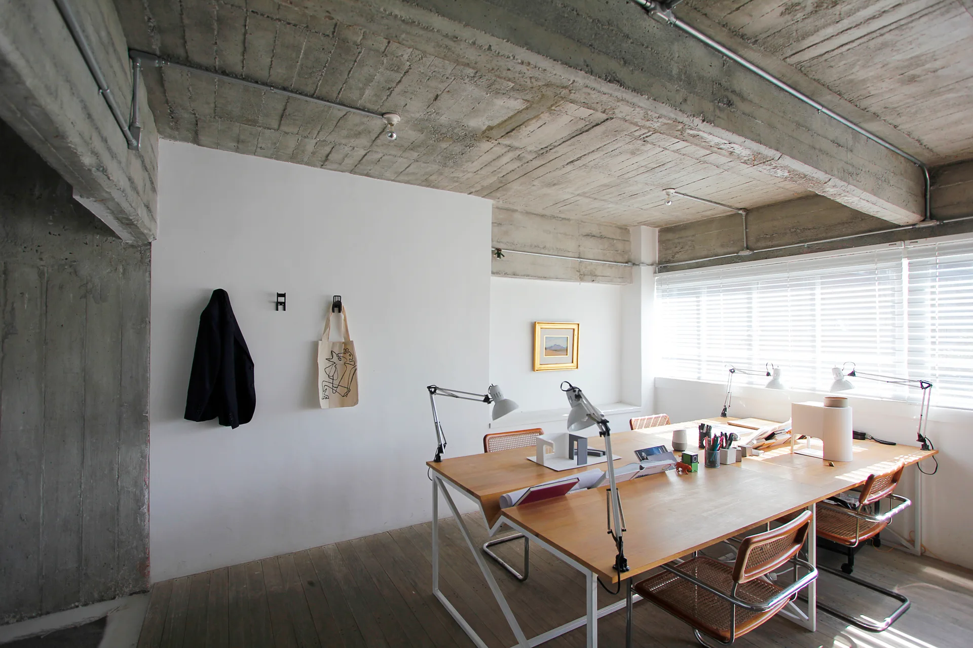 A minimalist studio features a large wooden desk paired with cane chairs beneath an exposed raw concrete ceiling with metal conduit piping.