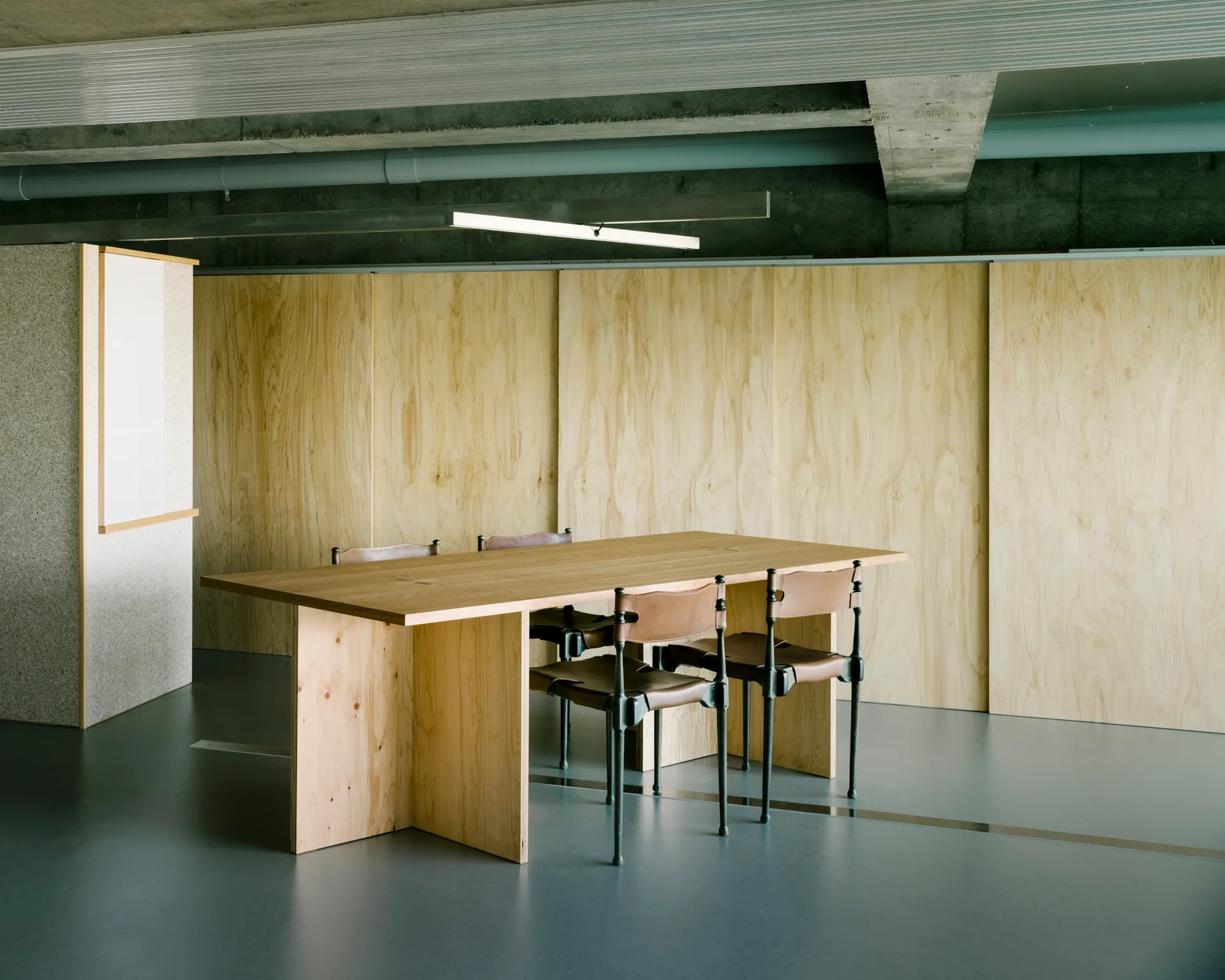 A light wood dining table with leather chairs stands in a room with warm plywood paneled walls and an exposed concrete ceiling.