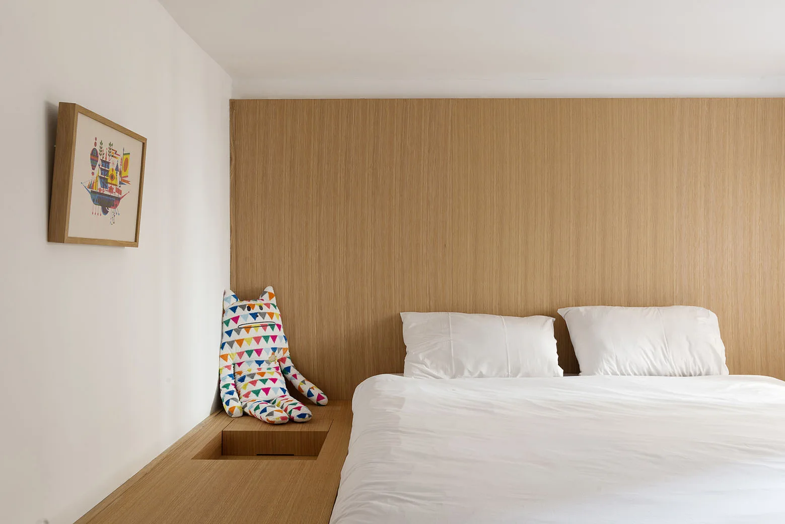 A minimalist bedroom features a bed with white linens on a light wood platform next to a matching wood-paneled wall.