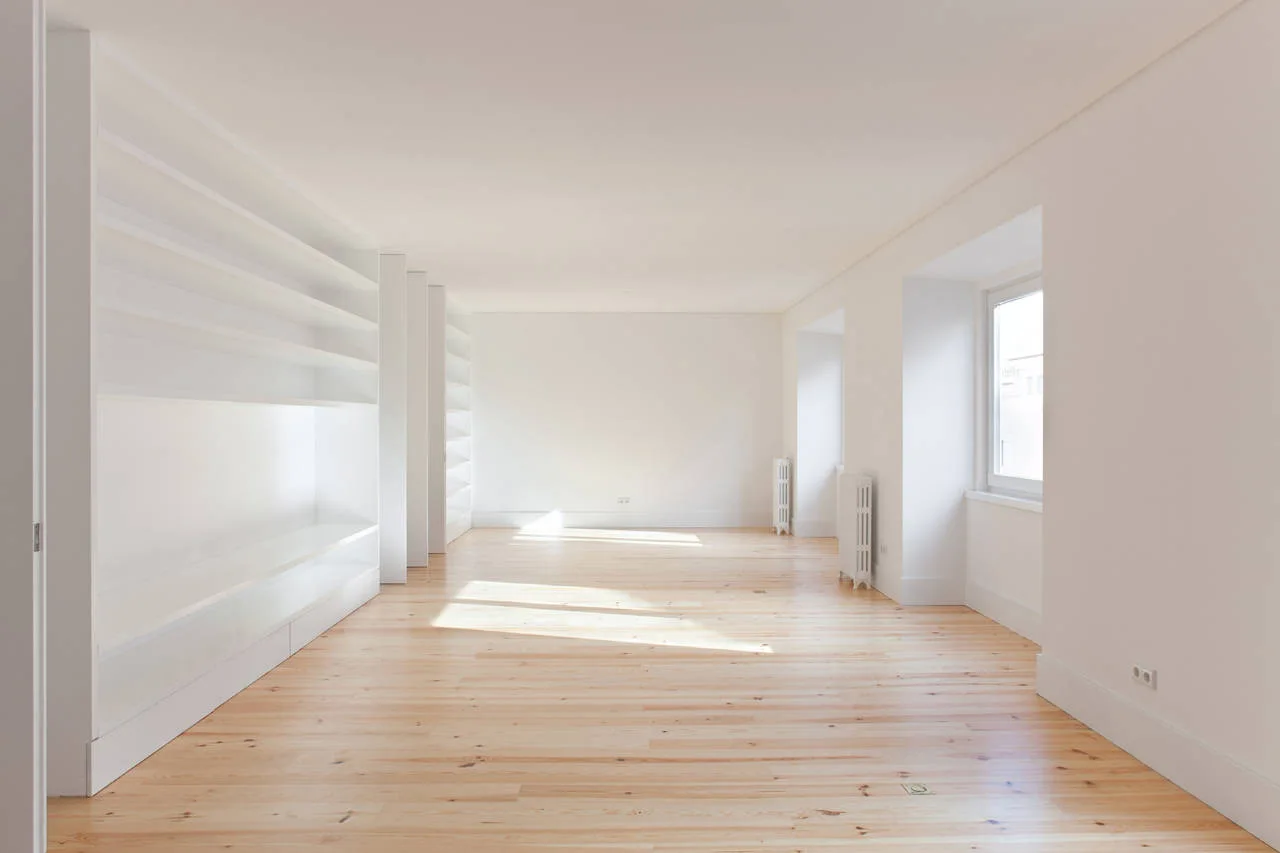 An empty minimalist room features natural light wood plank floors, smooth white walls, deep recessed windows, and a large white built-in shelving unit.