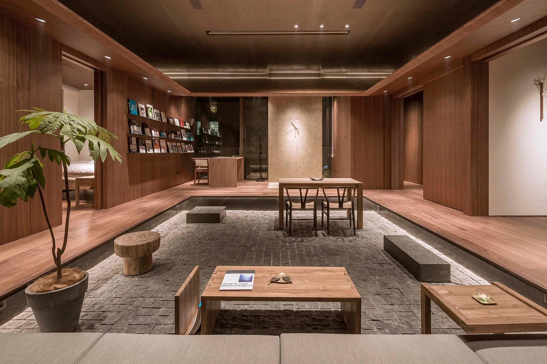 A sunken living room features a grey stone paved floor surrounded by raised wood platforms, wooden wall paneling, and warm recessed lighting.