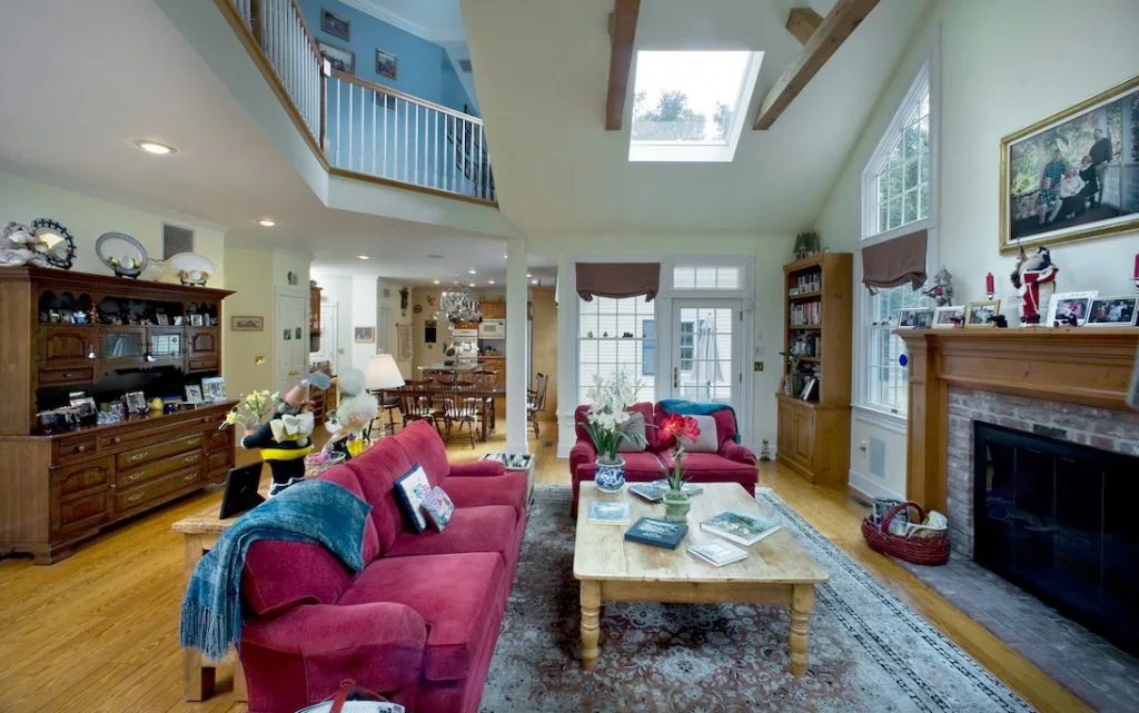 A vaulted ceiling living room featuring two red sofas, a light wood coffee table, and a brick fireplace under exposed wooden beams.