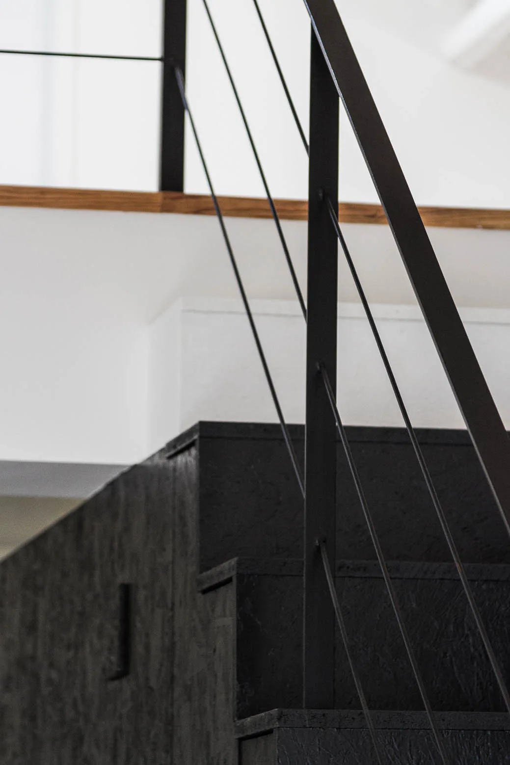 A close-up of a modern black staircase made of textured oriented strand board with a slim black metal cable railing against plain white walls.