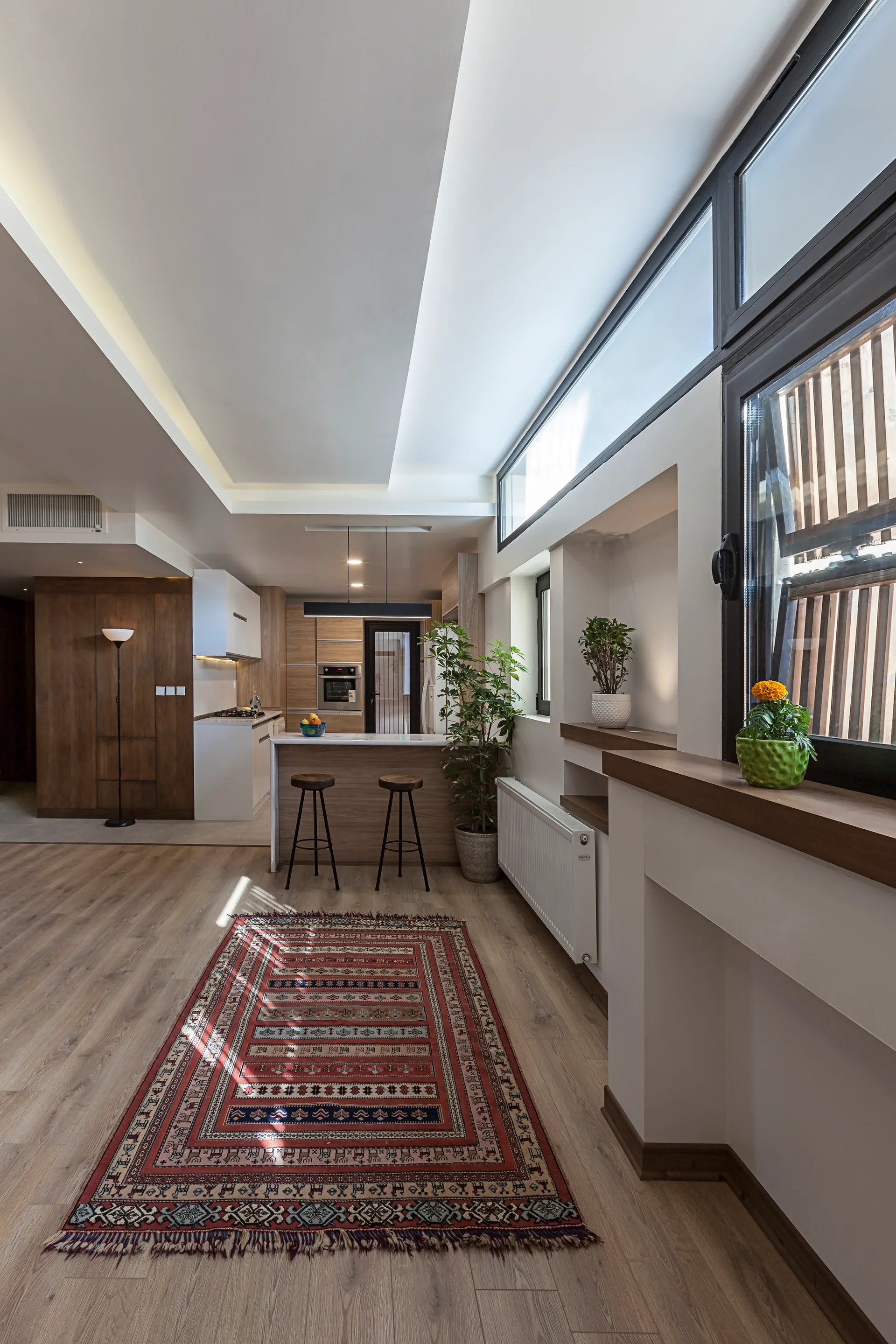 A modern open kitchen features a wooden island with bar stools, a red patterned runner rug on light wood floors, and high windows.