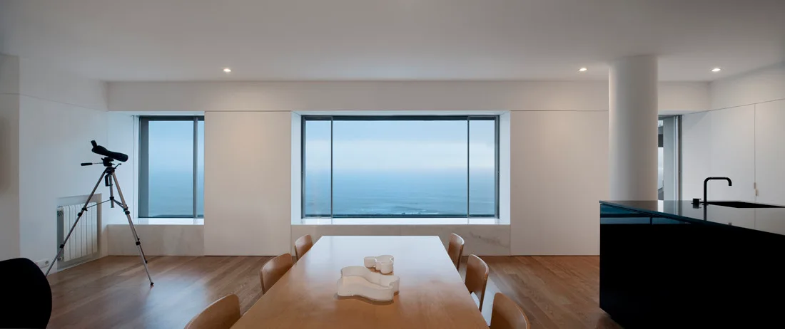 A minimalist dining room features a light wood table, chairs, a black kitchen island, and large picture windows overlooking the ocean.