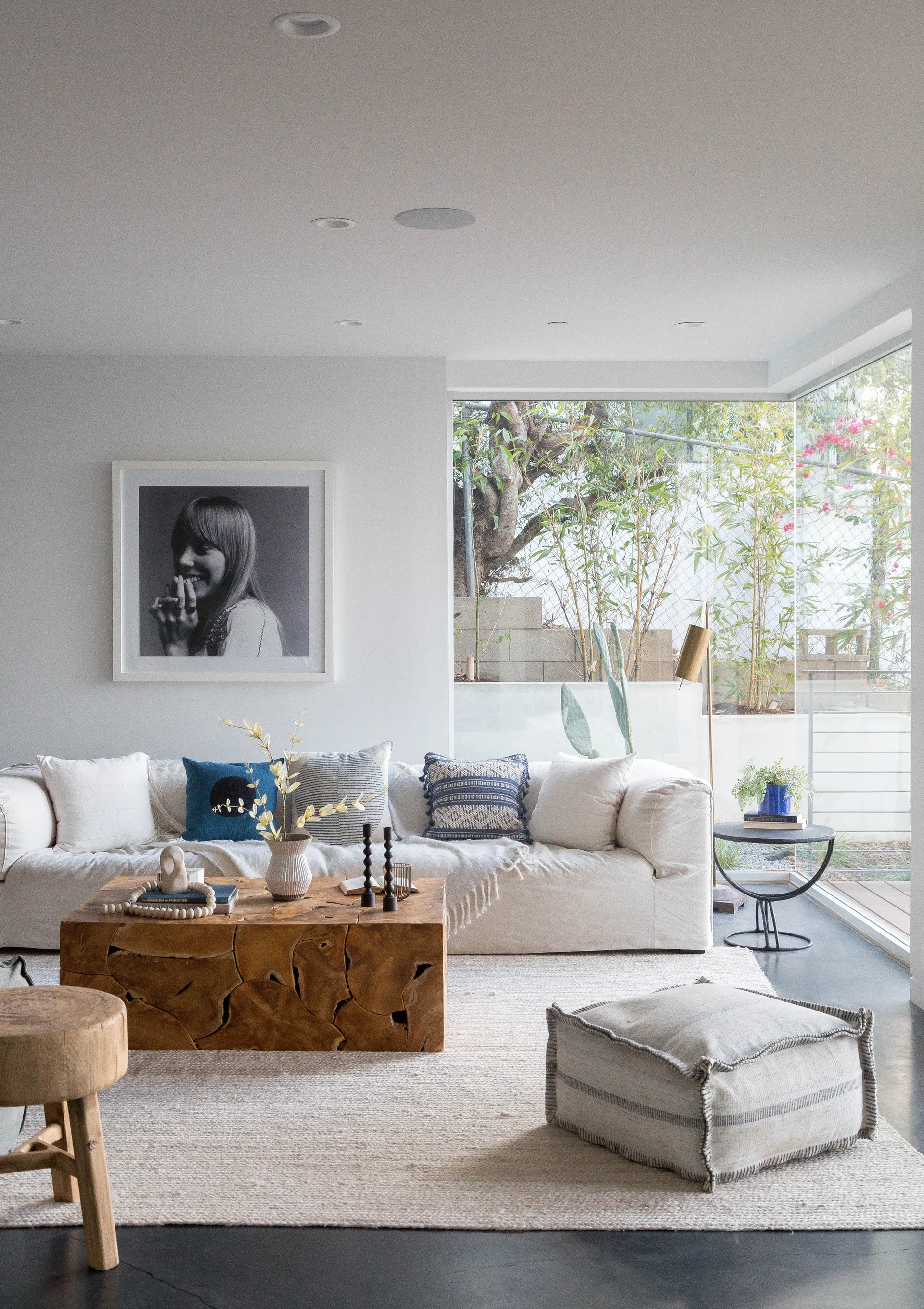 A bright living room features a cream-colored sofa, a solid wood block coffee table, a textured rug, and a large corner window showing garden greenery.