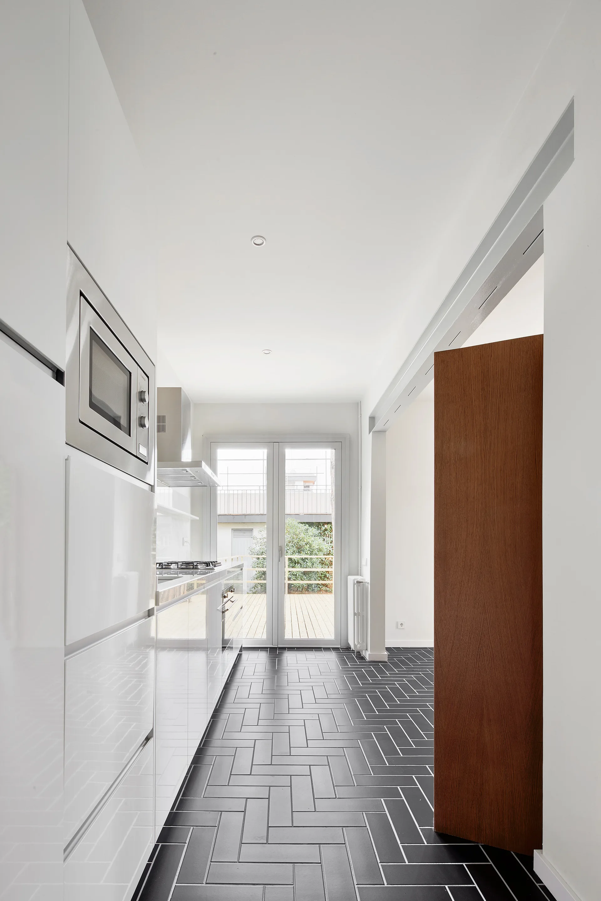 A narrow white kitchen features glossy handleless cabinets, a built-in microwave, and black herringbone-patterned floor tiles leading to glass doors.