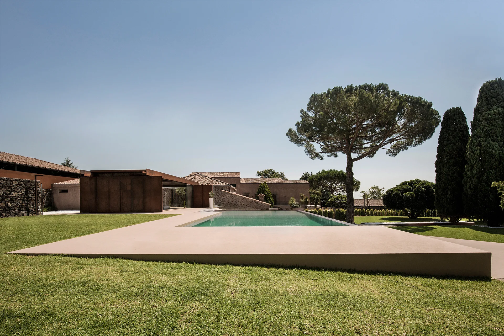 A modern rectangular swimming pool with a smooth concrete deck is surrounded by a green lawn, mature trees, and a house with rusted metal siding.