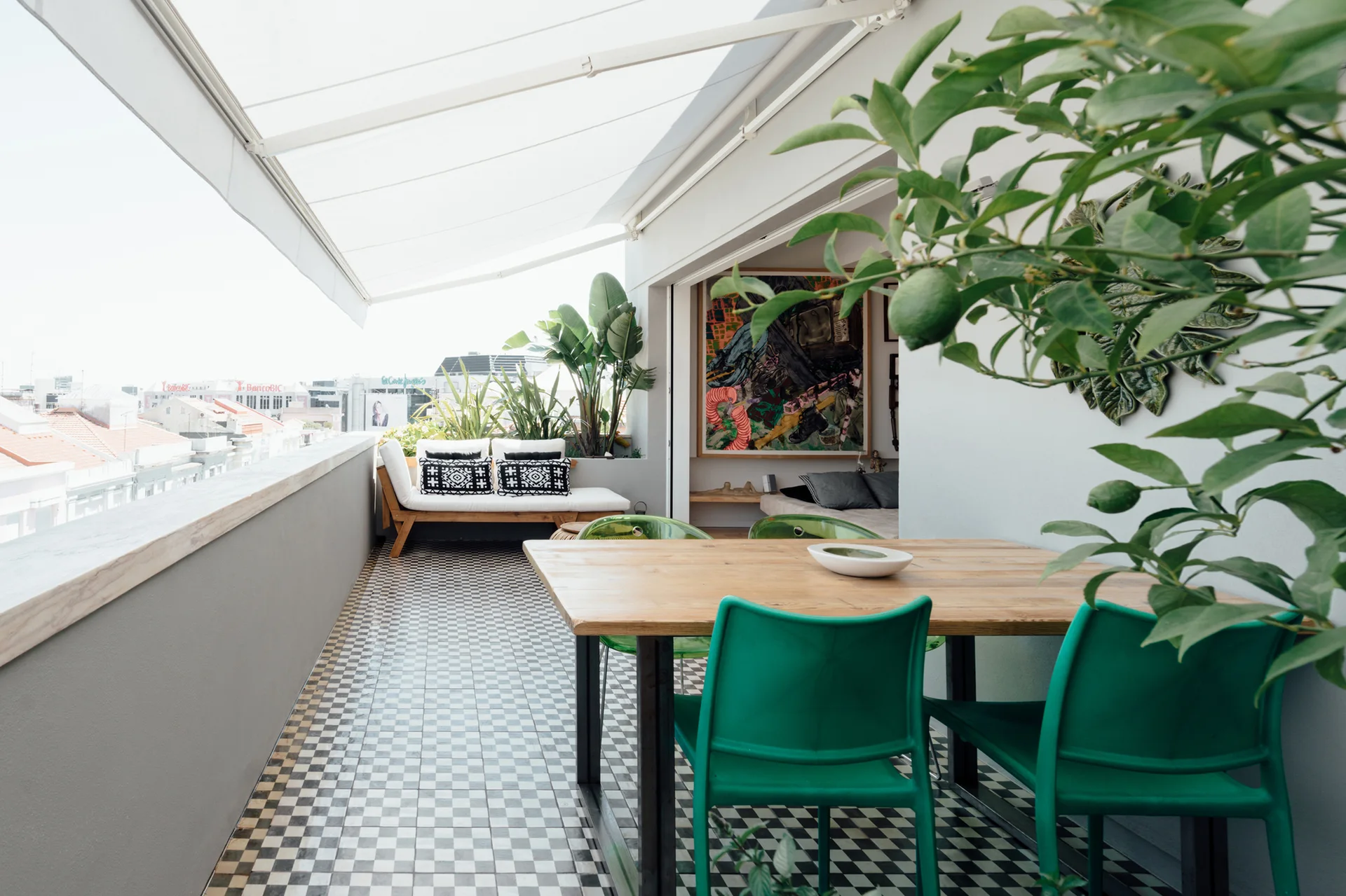 An outdoor balcony features a black and white checkered tile floor, a wooden dining table with green chairs, and a daybed under a white awning.