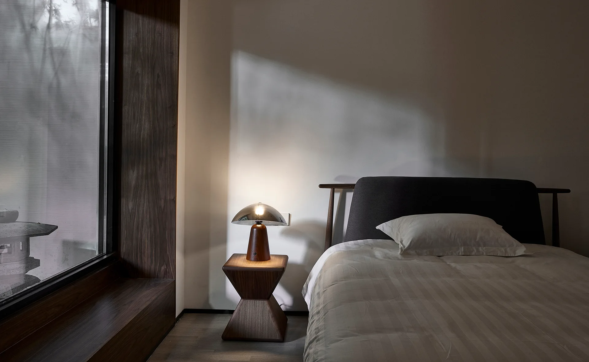 A minimalist bedroom features a bed with a dark headboard next to a sculptural wooden nightstand, a metal dome lamp, and a large deep-set window.