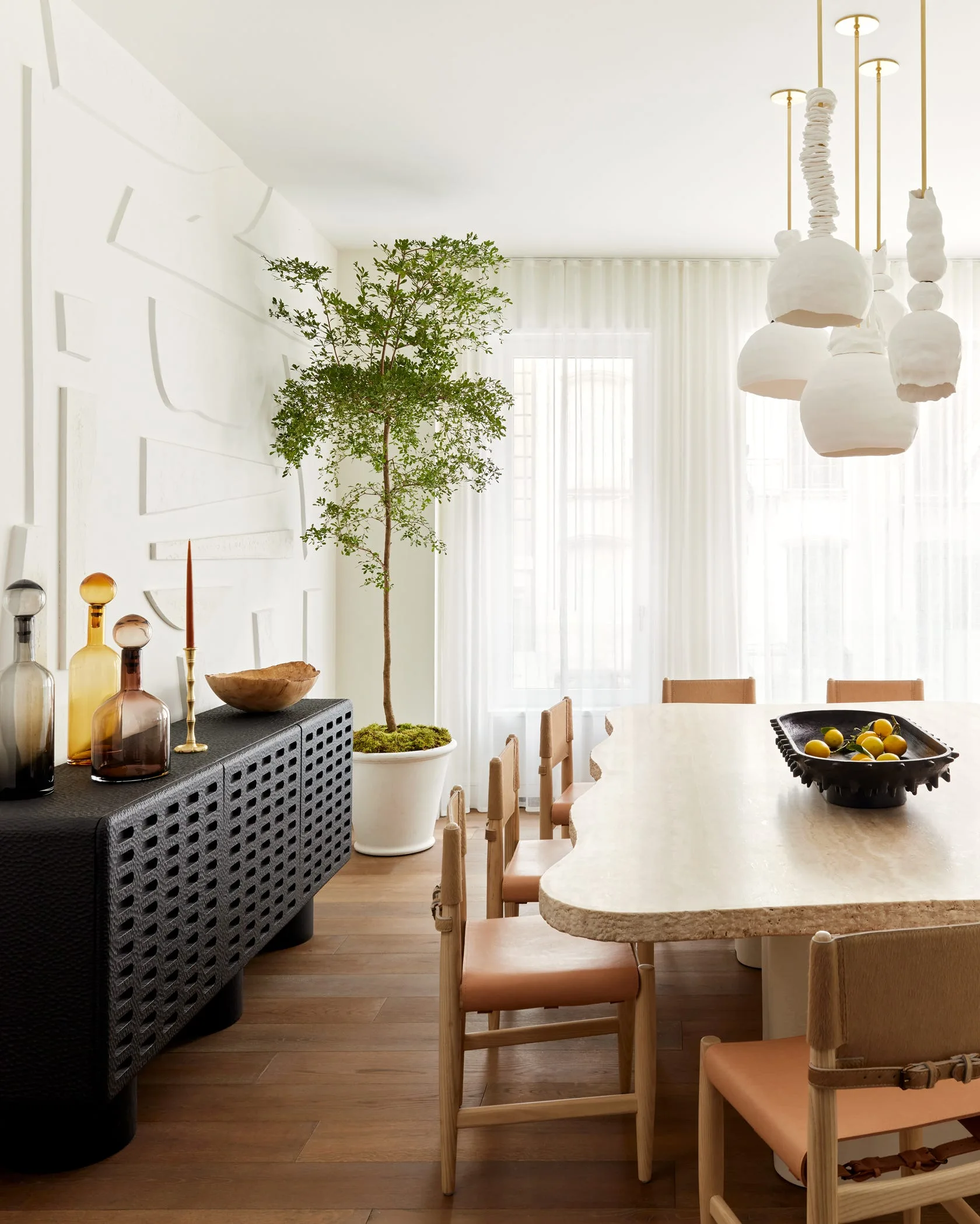 A bright dining room features a textured stone live-edge table, wood and leather chairs, a black patterned sideboard, and sculptural white pendant lights.