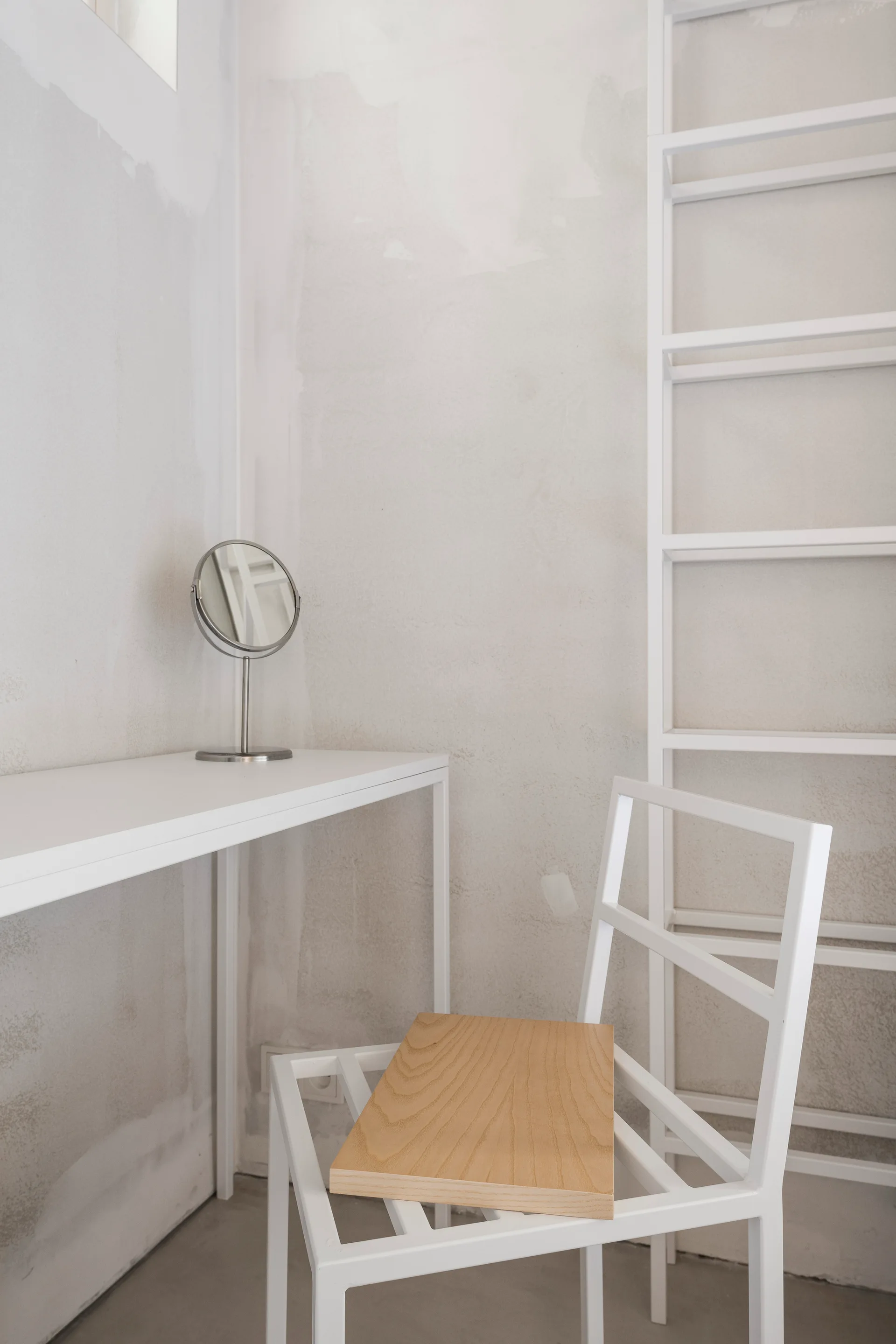 A minimalist white metal console table and chair with a wood seat stand against a textured raw plaster wall in a modern dressing area.