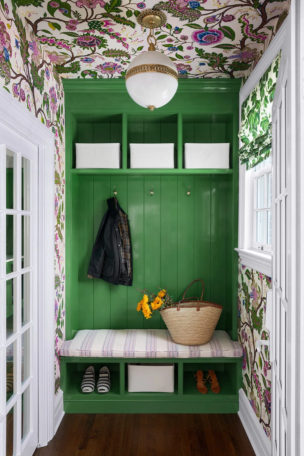 A mudroom features a built-in green storage bench and shelving unit surrounded by colorful floral wallpaper on the walls and ceiling.