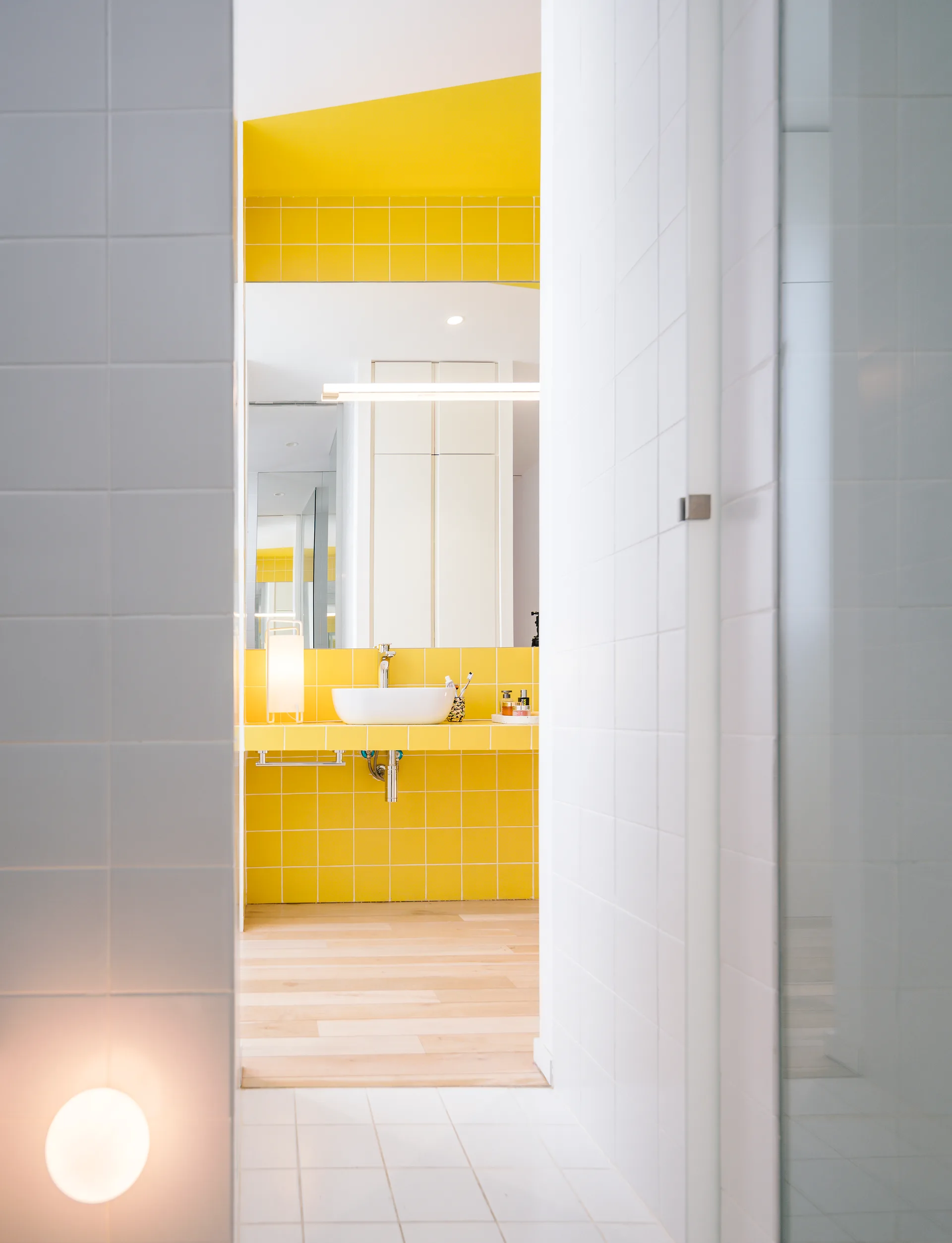 A peek through a white-tiled entryway into a bathroom with a bright yellow tiled vanity, a white vessel sink, and light wood floors.
