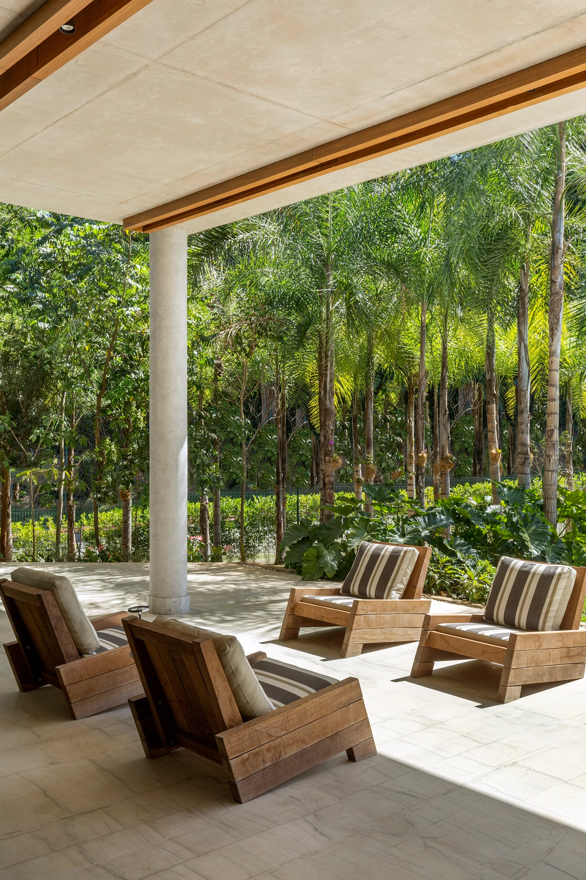 Four wooden lounge chairs with striped cushions sit on a light stone patio beneath a concrete overhang, facing a lush tropical garden.
