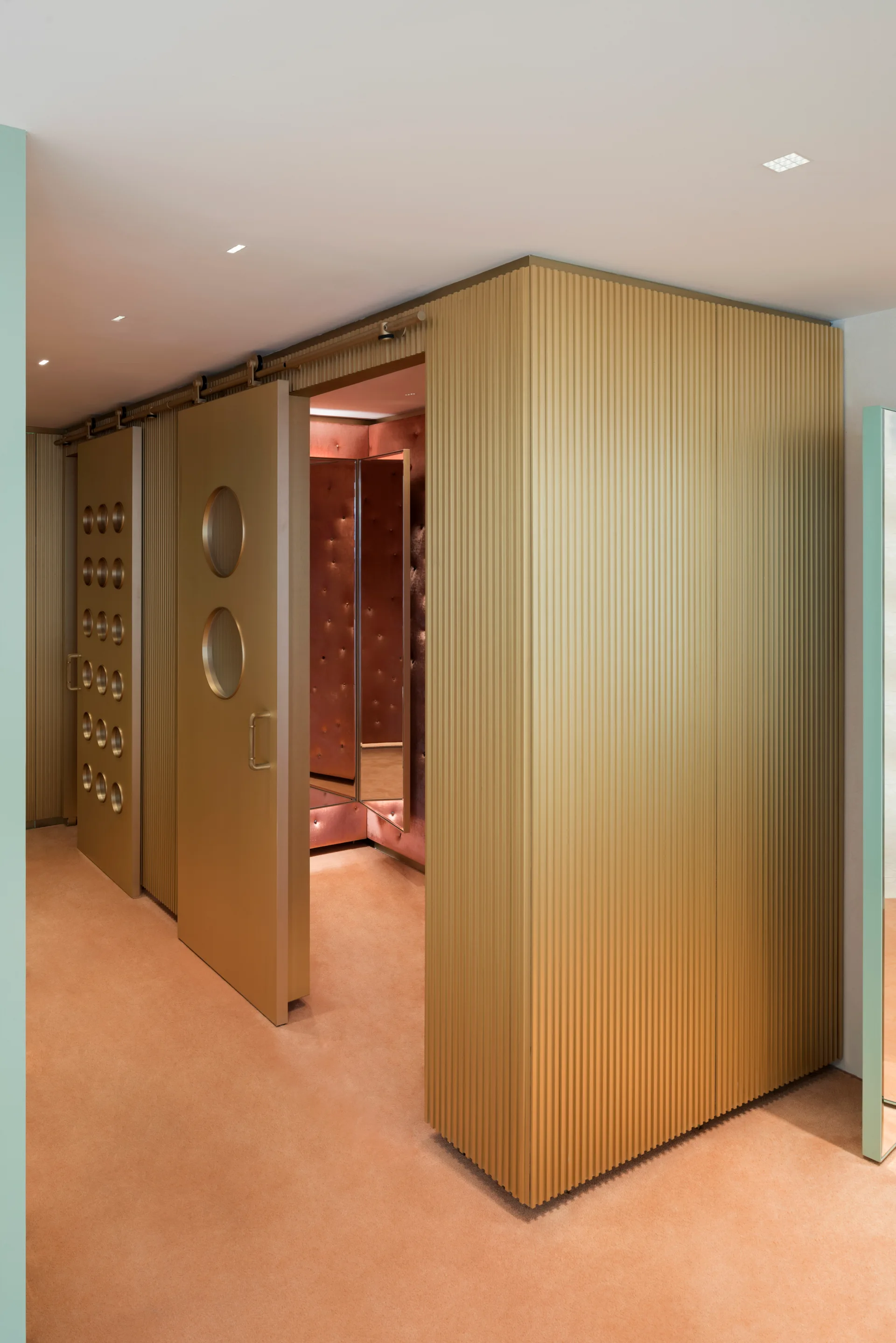 A modern dressing room area featuring gold ribbed wall panels, metallic sliding doors with circular portholes, and peach-colored carpet.