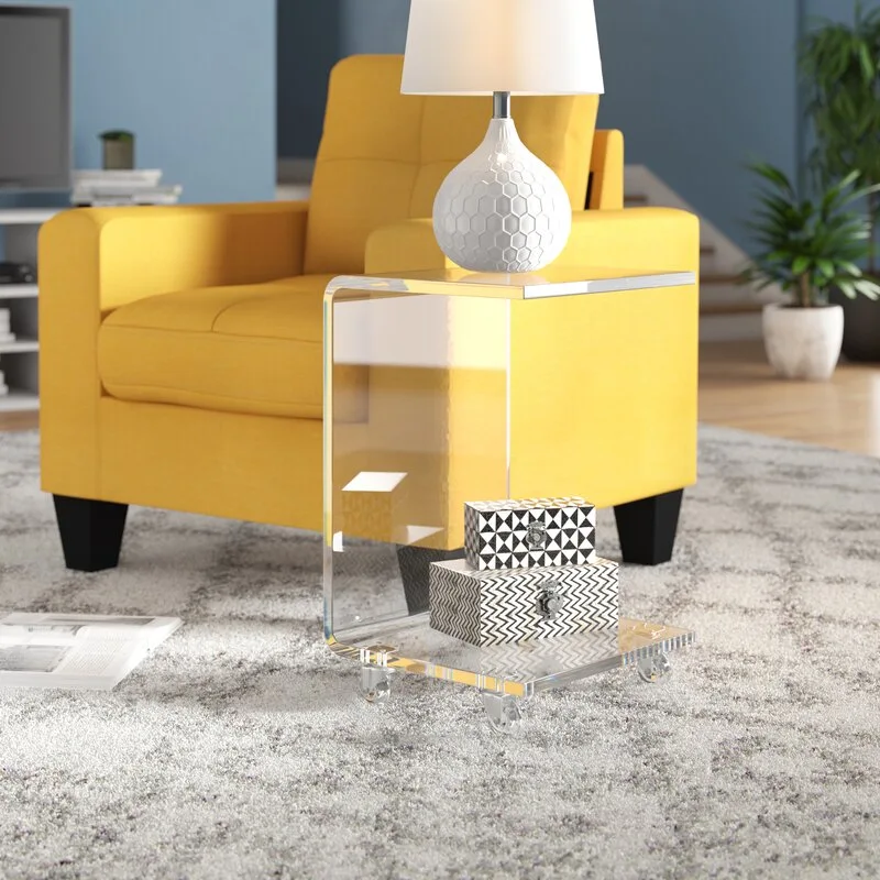 A clear acrylic rolling end table sits on a patterned grey rug next to a bright yellow armchair and features a white table lamp.