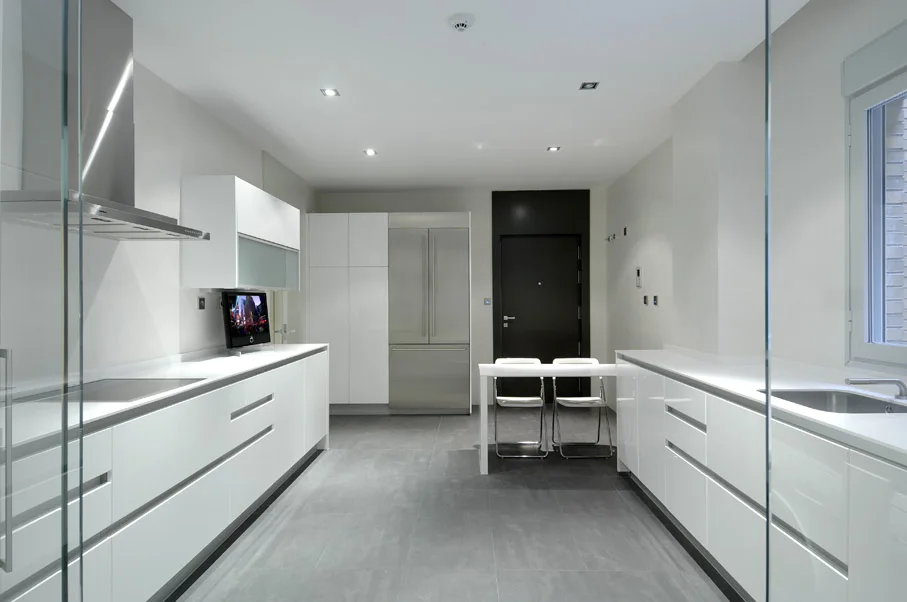A minimalist galley kitchen featuring glossy white cabinets, grey tile flooring, stainless steel appliances, and a small white dining table in the back.