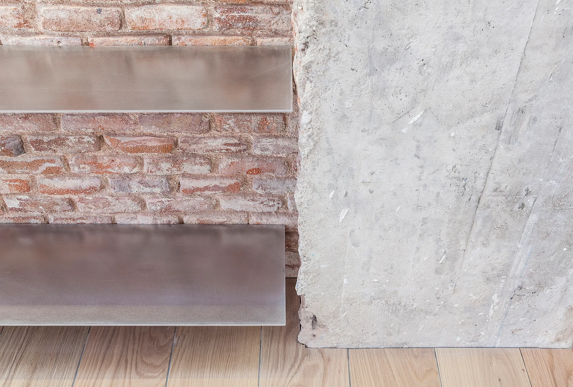 Two floating metal shelves are mounted on an exposed red brick wall next to a rough grey concrete column above light wood flooring.