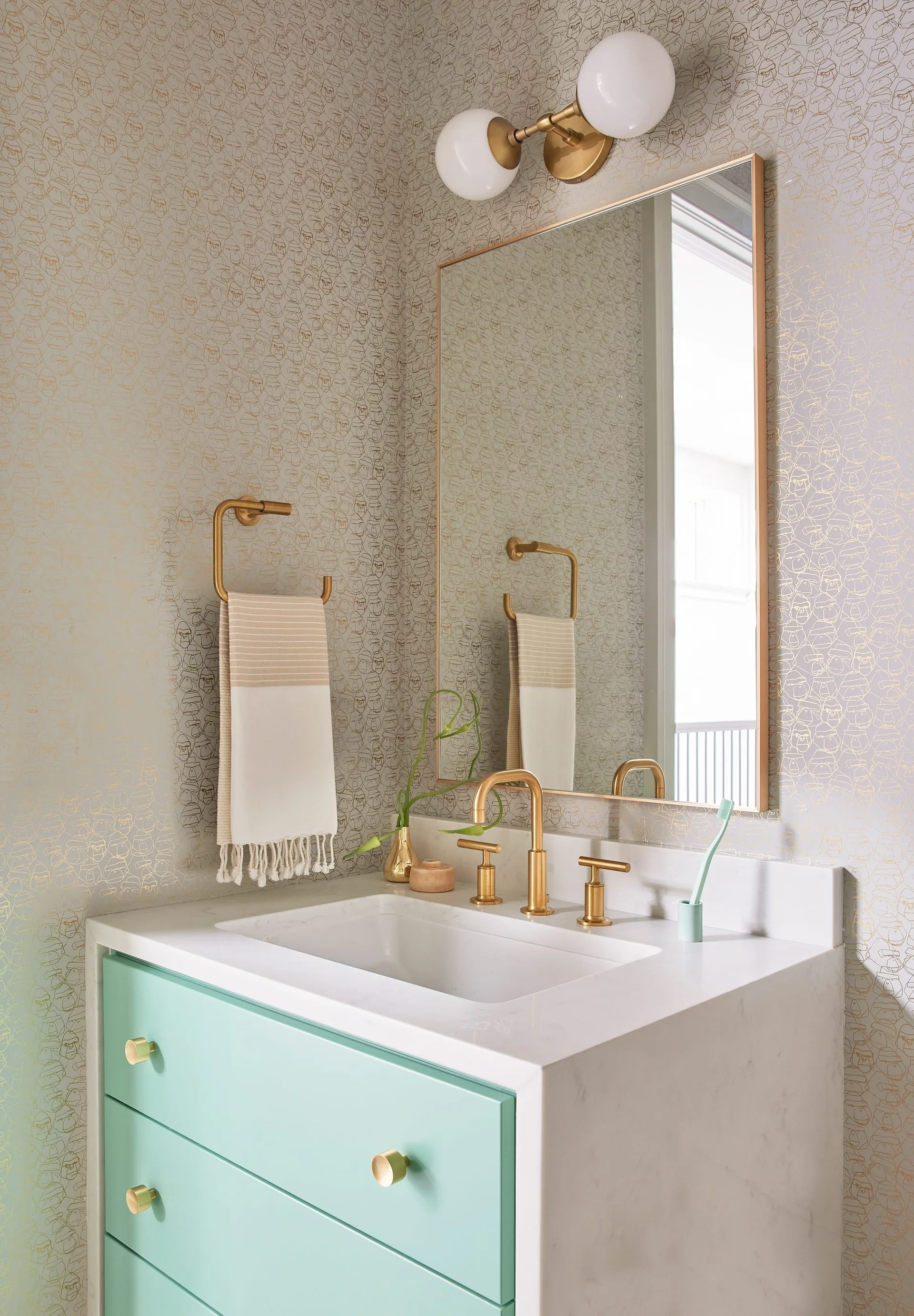 A powder room features a mint green vanity with a stone countertop, brushed gold faucet, rectangular mirror, and patterned metallic wallpaper.