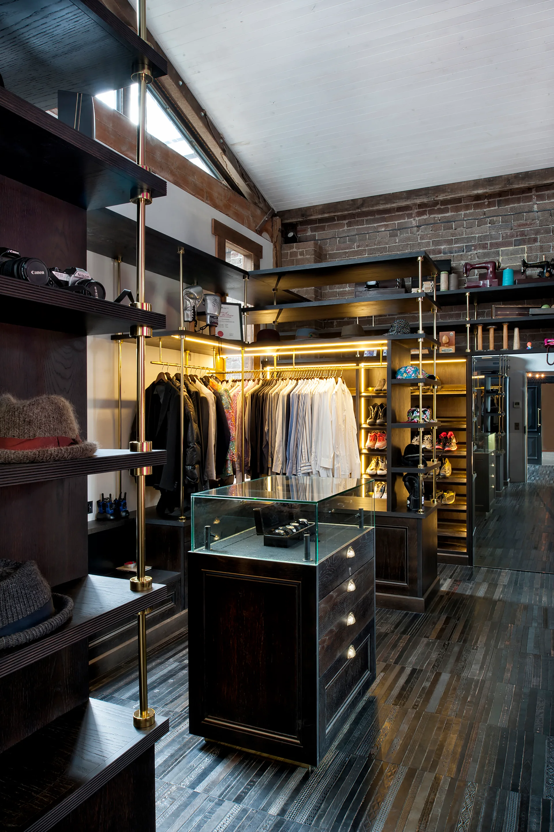 An industrial walk-in closet featuring dark wood shelving supported by brass poles, a central glass-topped drawer island, and an exposed brick wall.