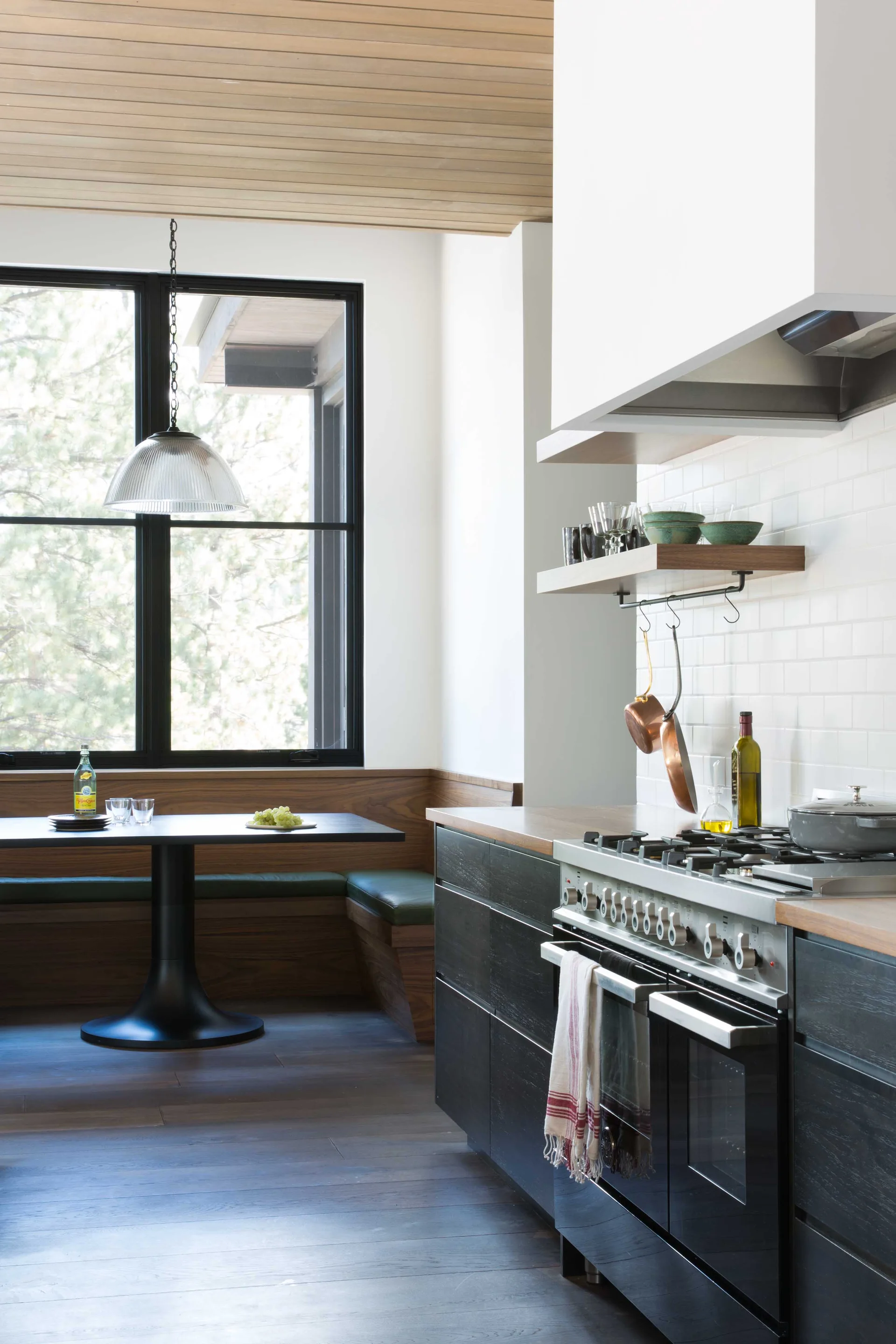 A modern kitchen features dark lower cabinets, a stainless steel range, and a built-in wooden corner banquette next to a black-framed window.