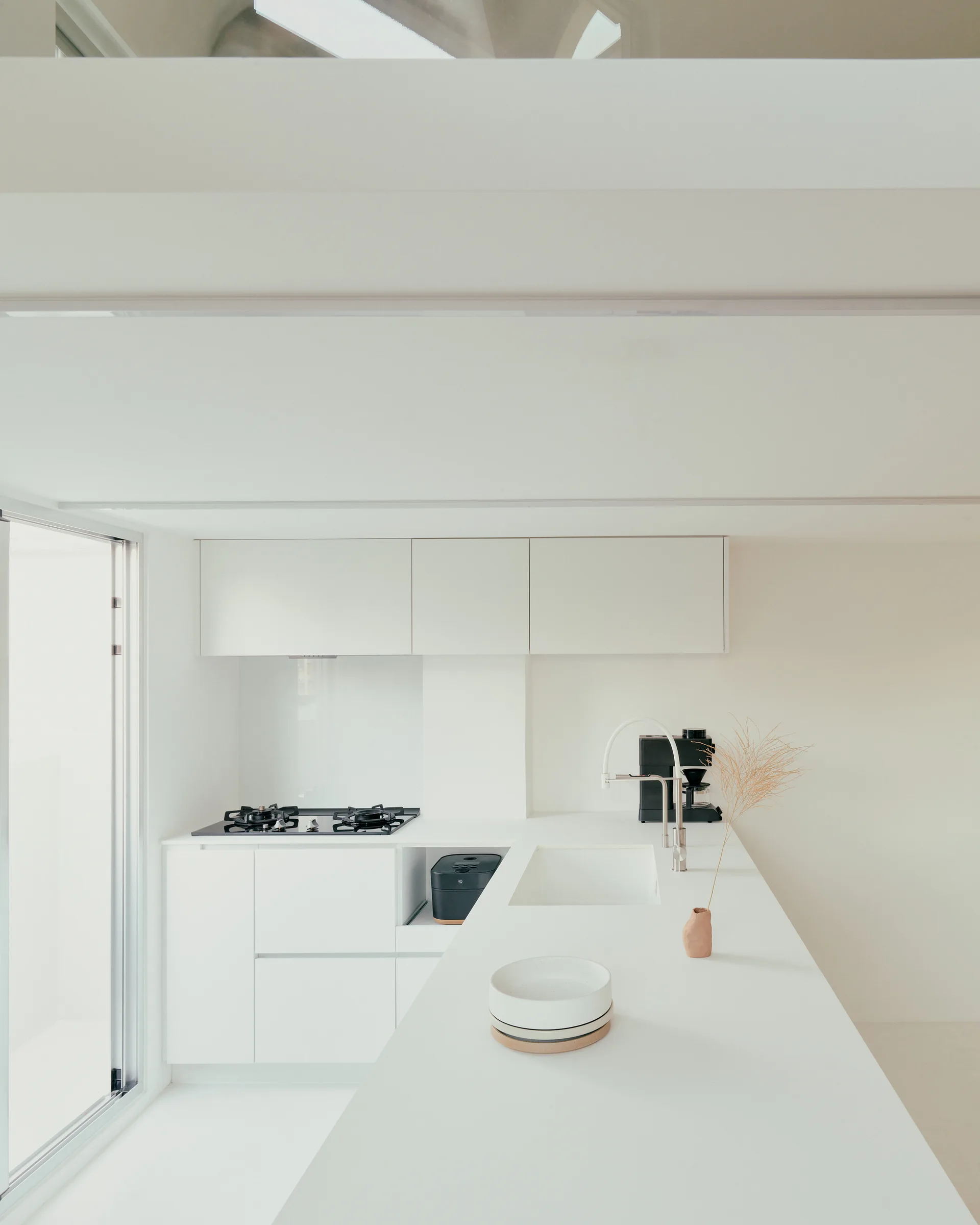 A minimalist white kitchen features a long central island with an integrated sink, handleless flat-panel cabinetry, and a black gas stovetop.