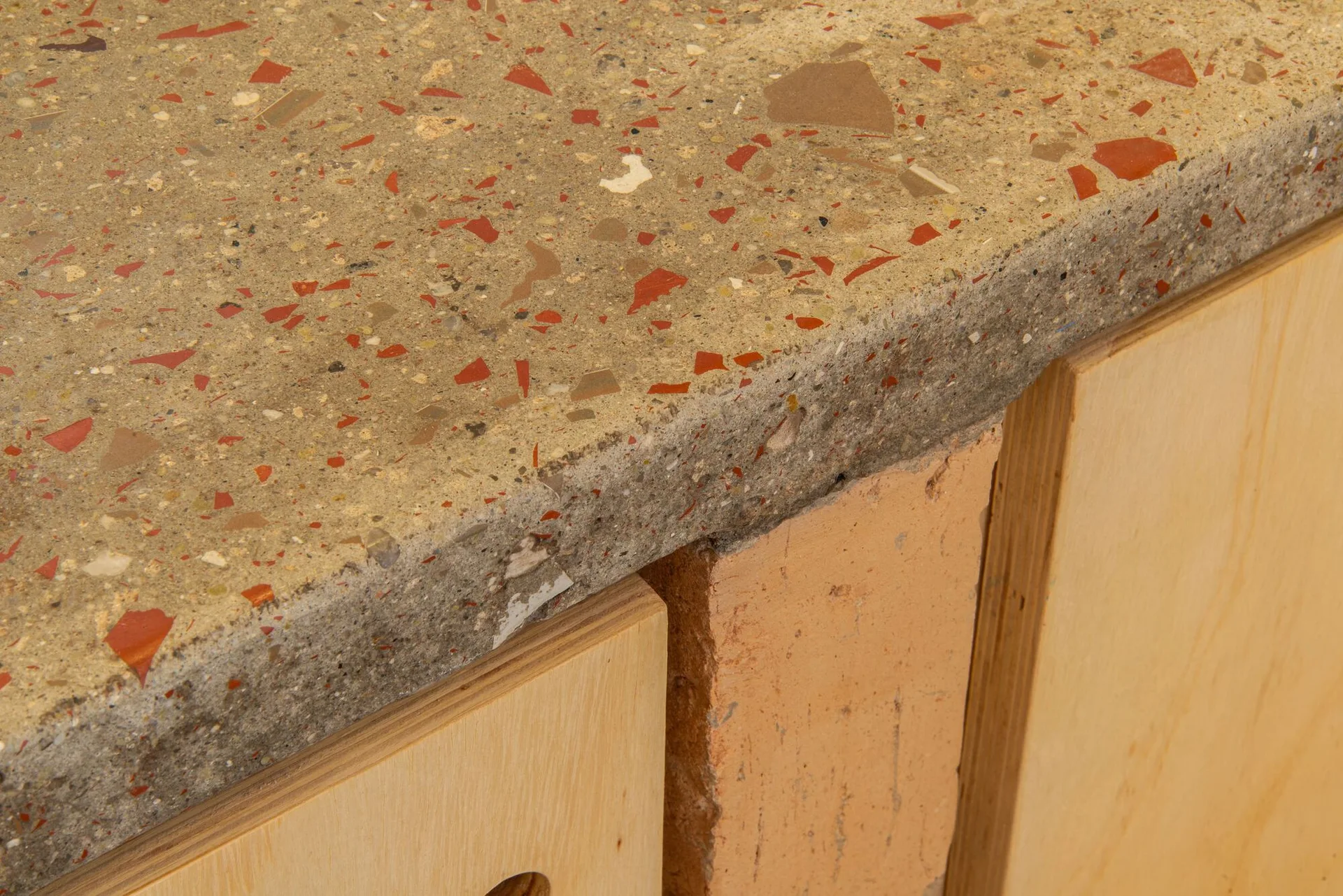 A close-up of a speckled terrazzo countertop with red and brown aggregates resting on light plywood cabinets and a raw brick support.