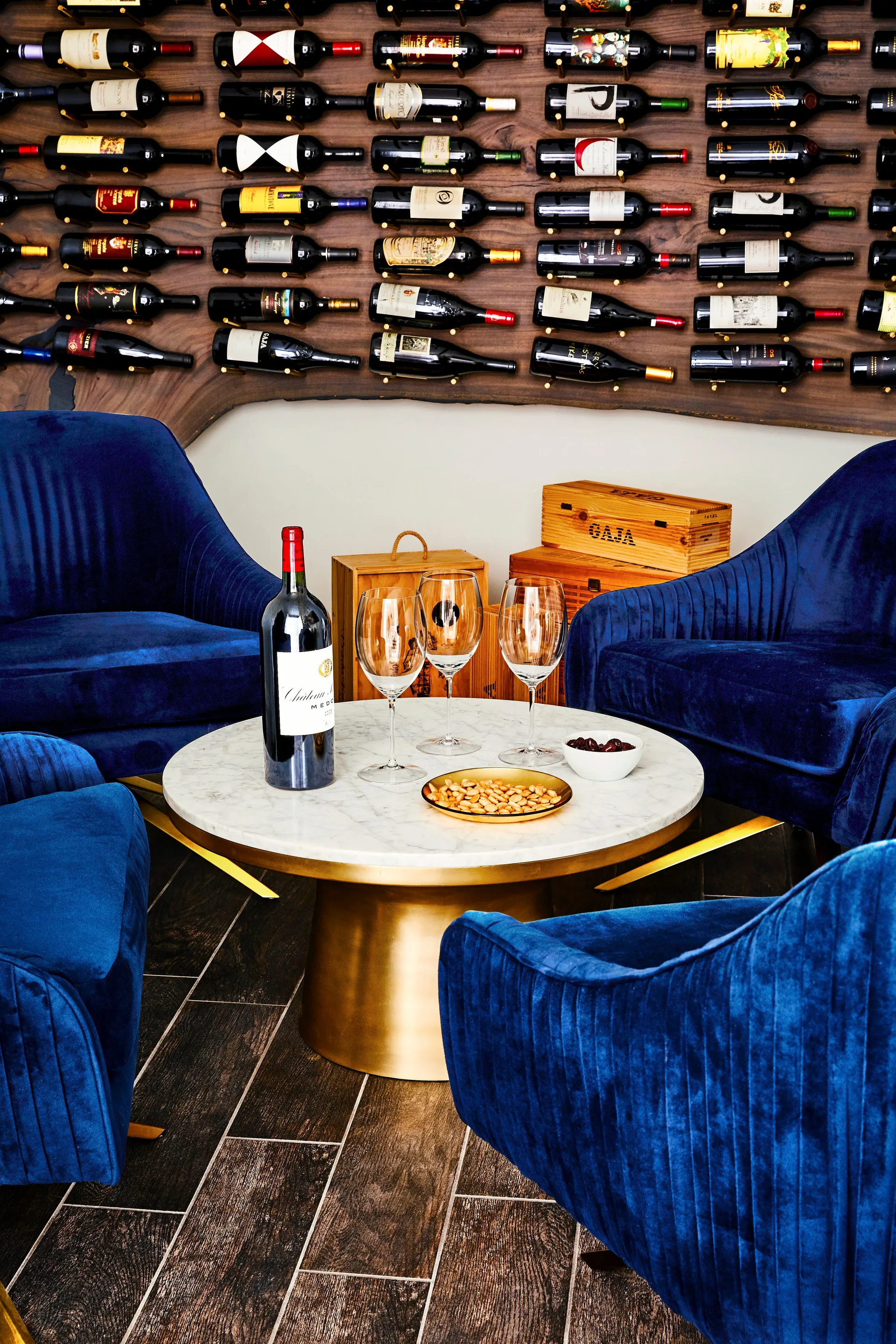 Deep blue velvet armchairs surround a round marble-topped coffee table with a brass base in front of a wall-mounted wine rack.