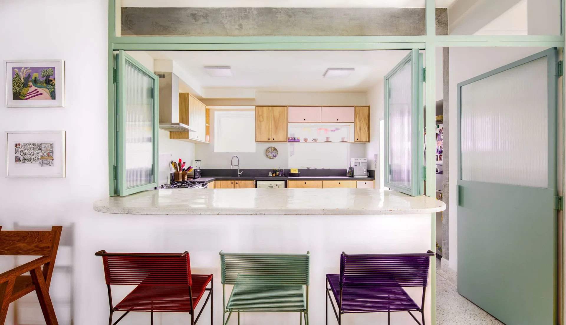 A kitchen pass-through with mint green trim and fluted glass windows features a stone-like counter with three colorful bar stools in red, green, and purple.