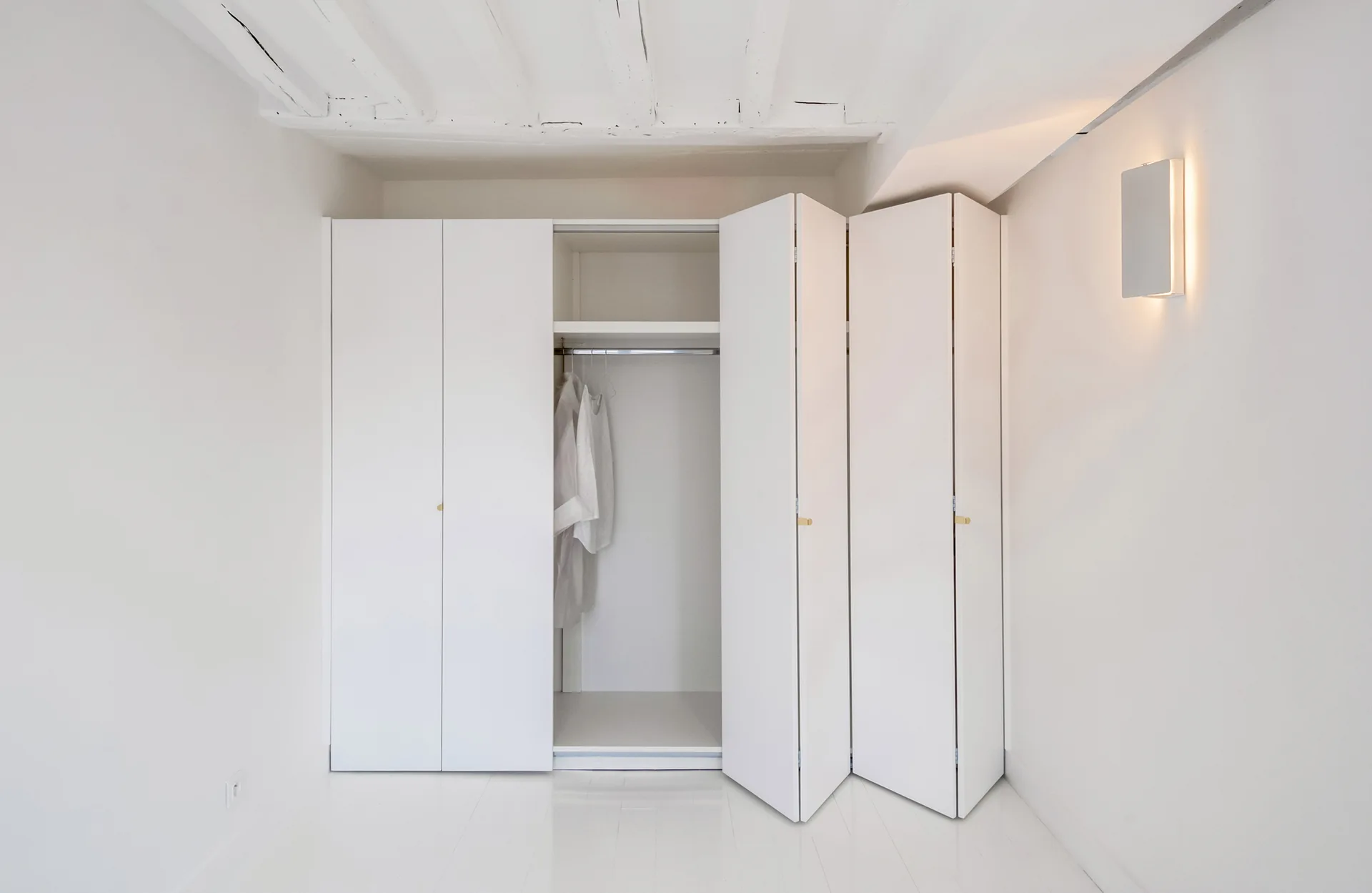 A minimalist white closet features open bi-fold doors, small gold hardware, and exposed ceiling beams in a room with a glowing wall sconce.
