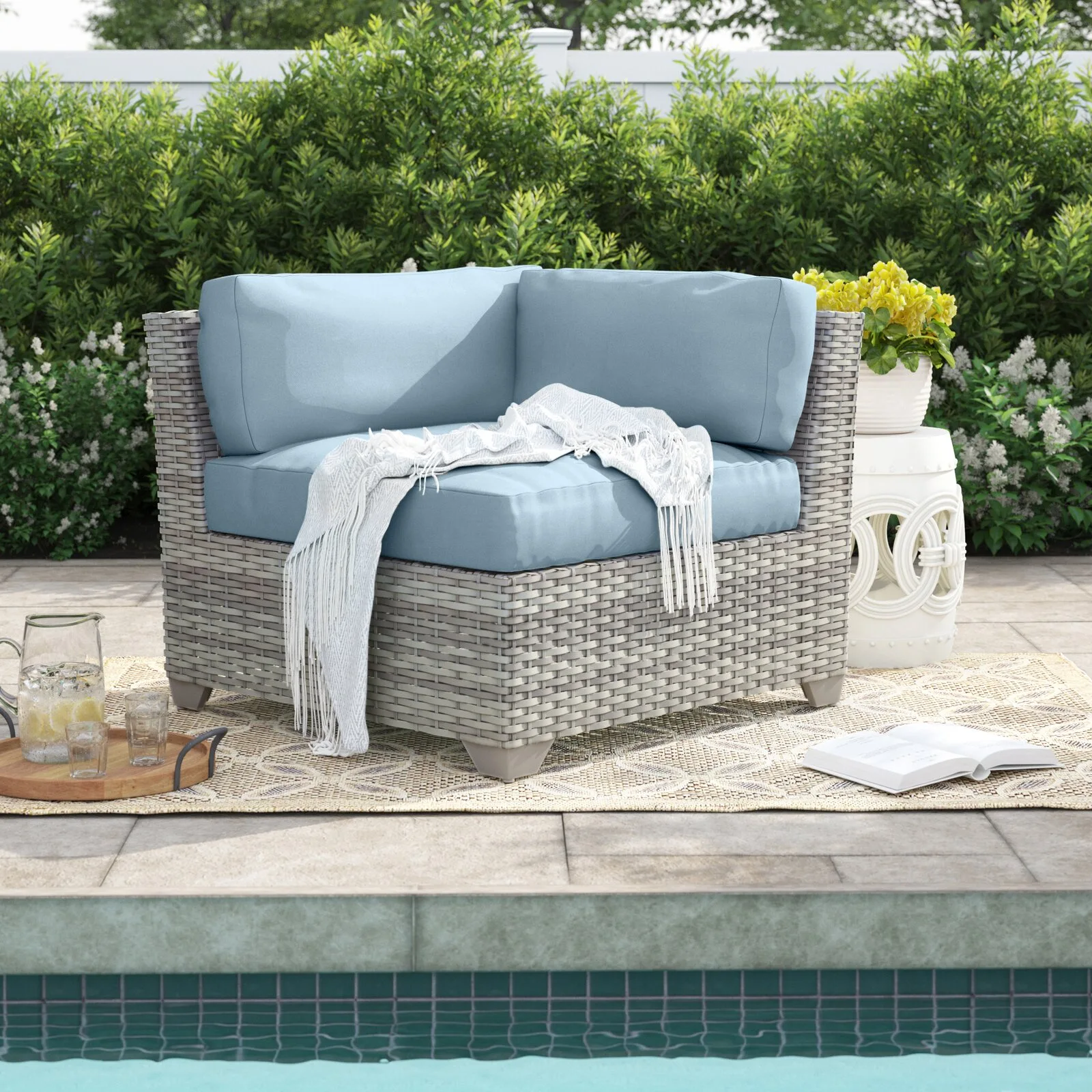 A grey woven wicker corner chair with light blue cushions sits on a patterned outdoor rug next to a white ceramic side table and swimming pool.