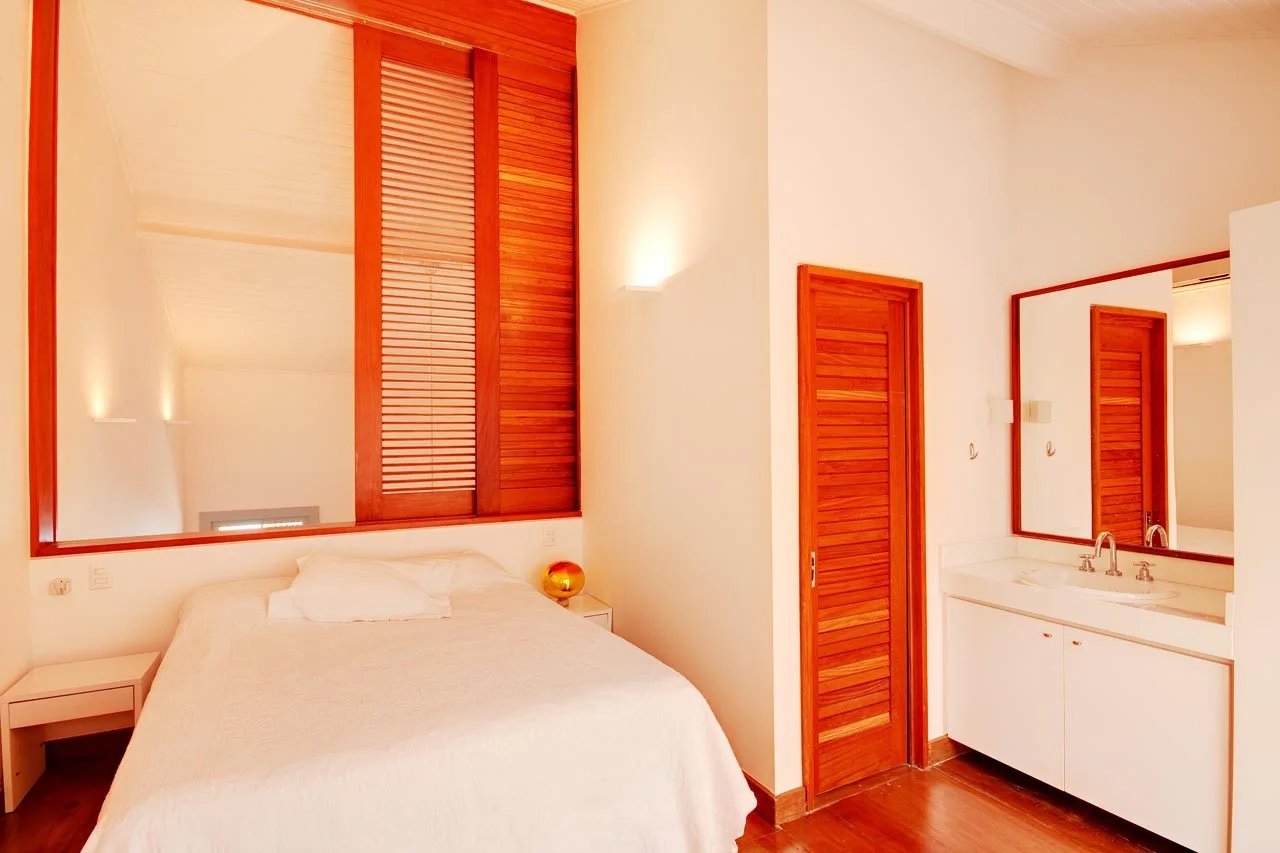 A minimalist bedroom features white bedding, an integrated bathroom vanity, warm wood floors, and a wood louvered door.