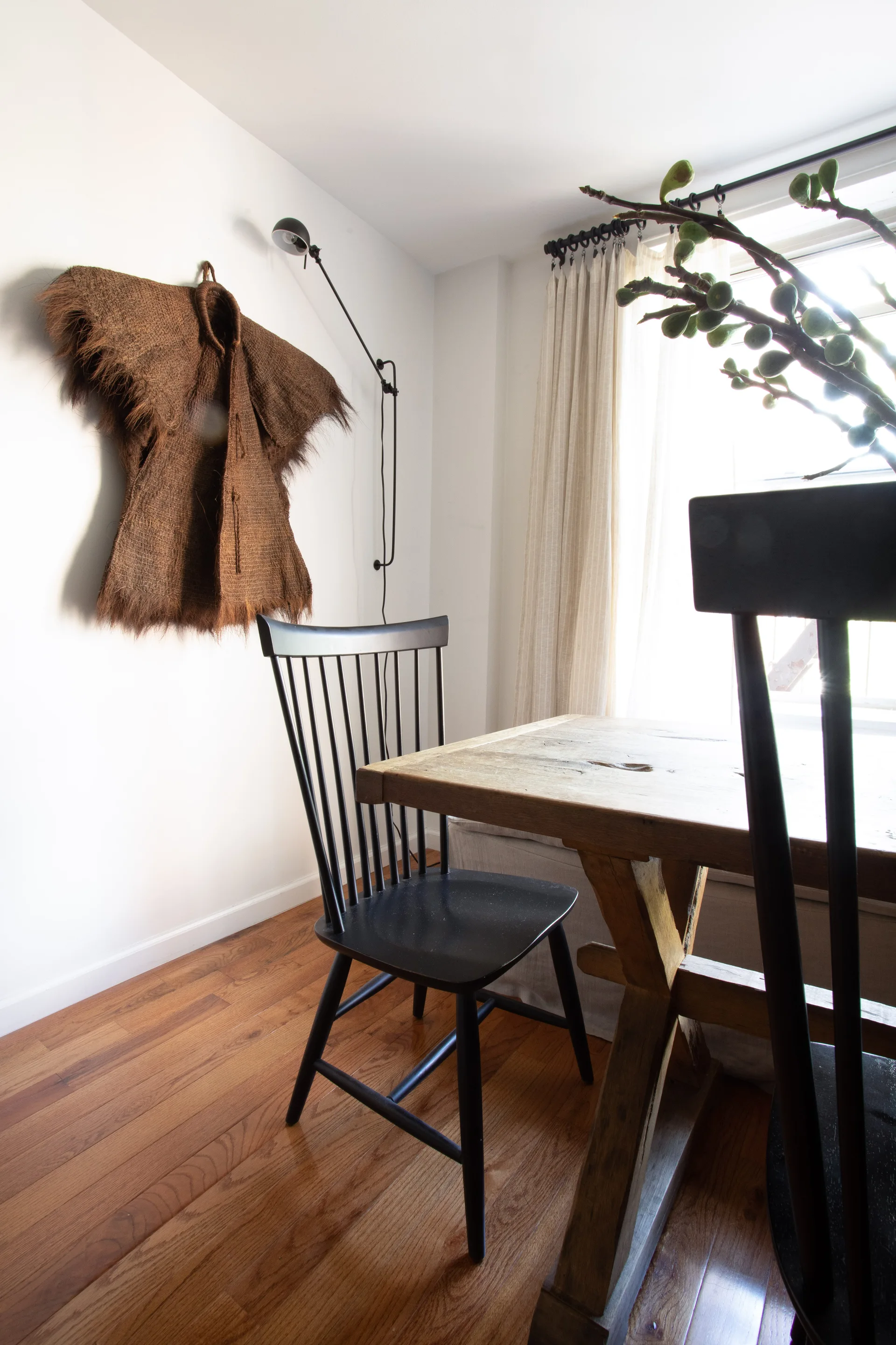 A bright dining area featuring a rustic wooden table with black spindle-back chairs, a textured fiber wall hanging, and a black wall-mounted sconce.