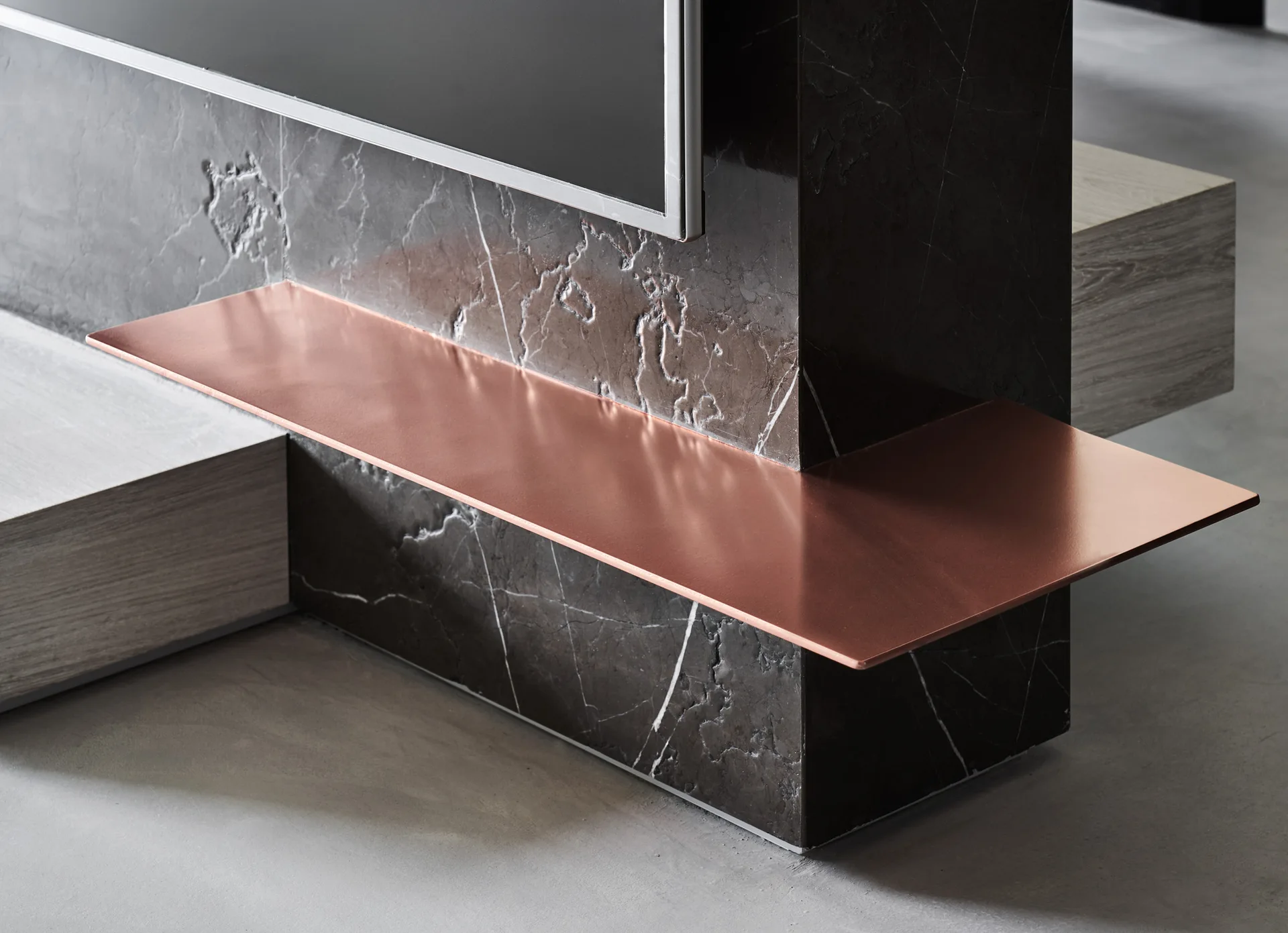 A close-up of a built-in media console featuring veined dark gray marble panels and a thin, sleek copper metal shelf.