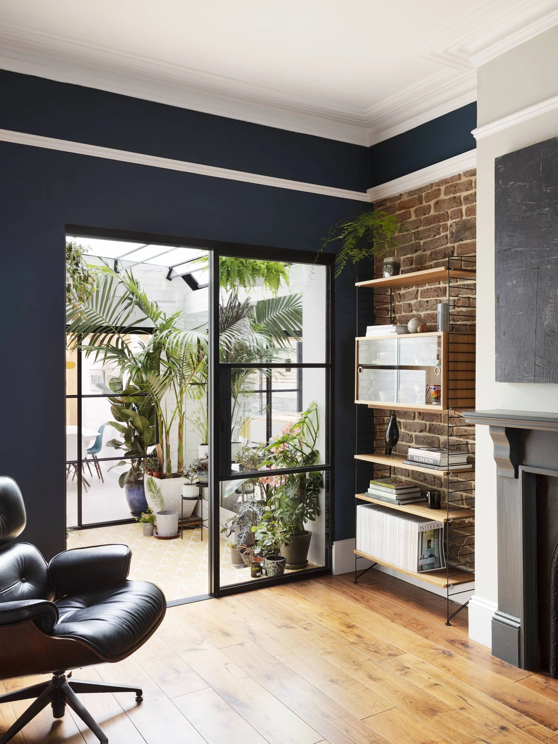 A living room with a black leather lounge chair, navy walls, and a metal-framed glass partition opening into a sunroom filled with potted plants.