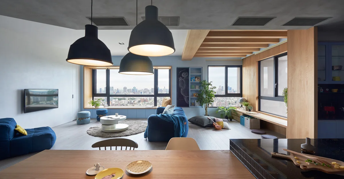 An open-concept living room features a blue sofa, a round tiered coffee table, a light blue built-in window bench, and exposed wood ceiling beams.