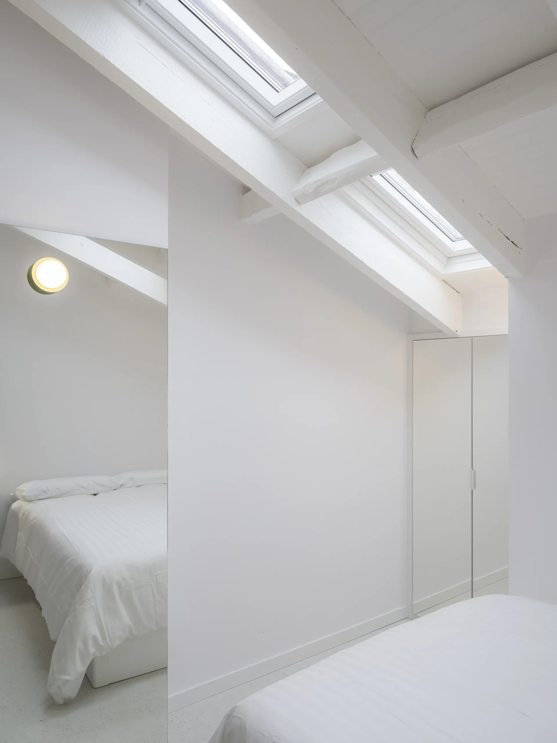 A minimalist white attic bedroom featuring a sloped ceiling with skylights, exposed white wooden beams, and a large floor-to-ceiling mirror.