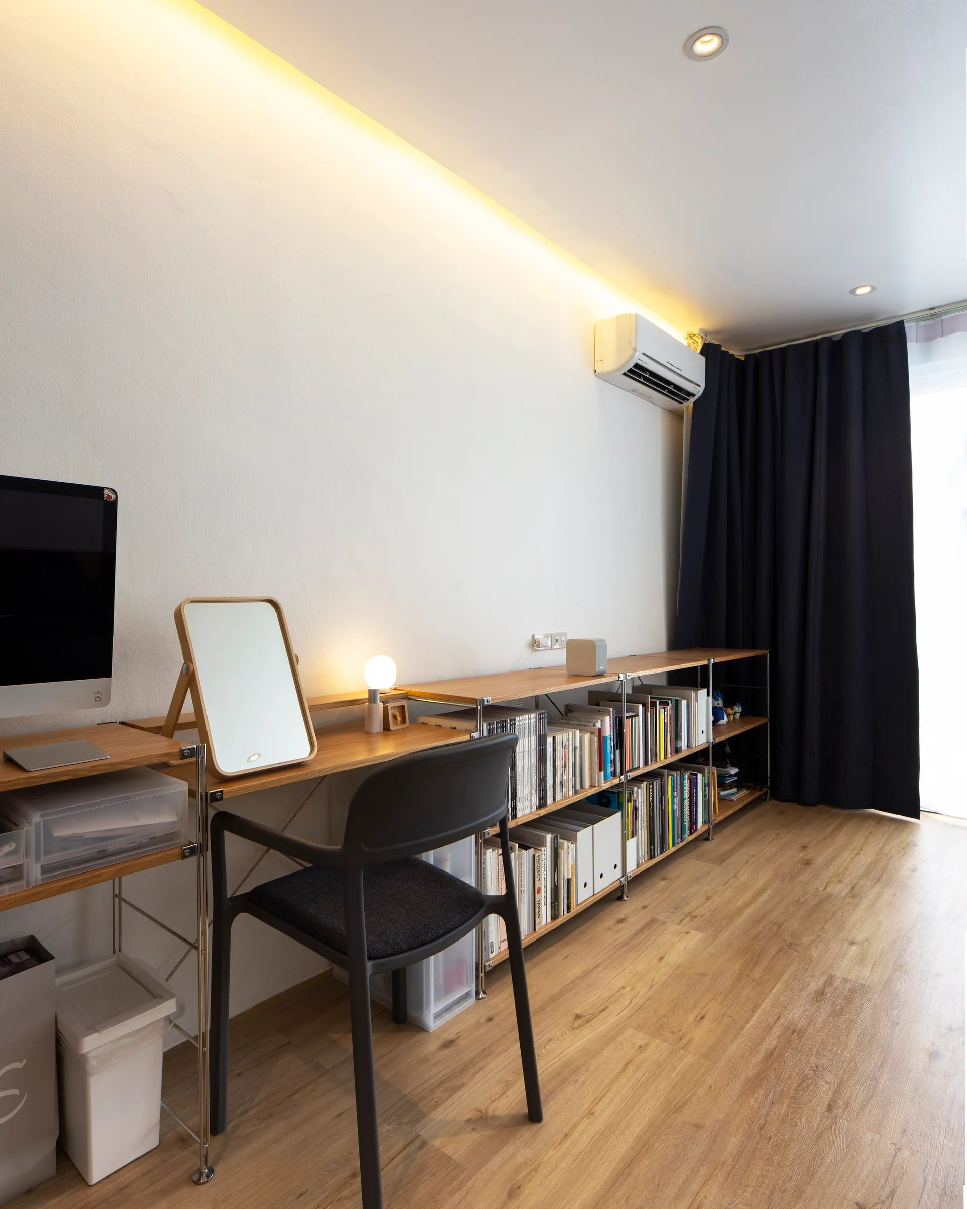 A minimalist home office features a long modular wooden desk with integrated shelving, a black chair, and soft yellow cove lighting along the white wall.