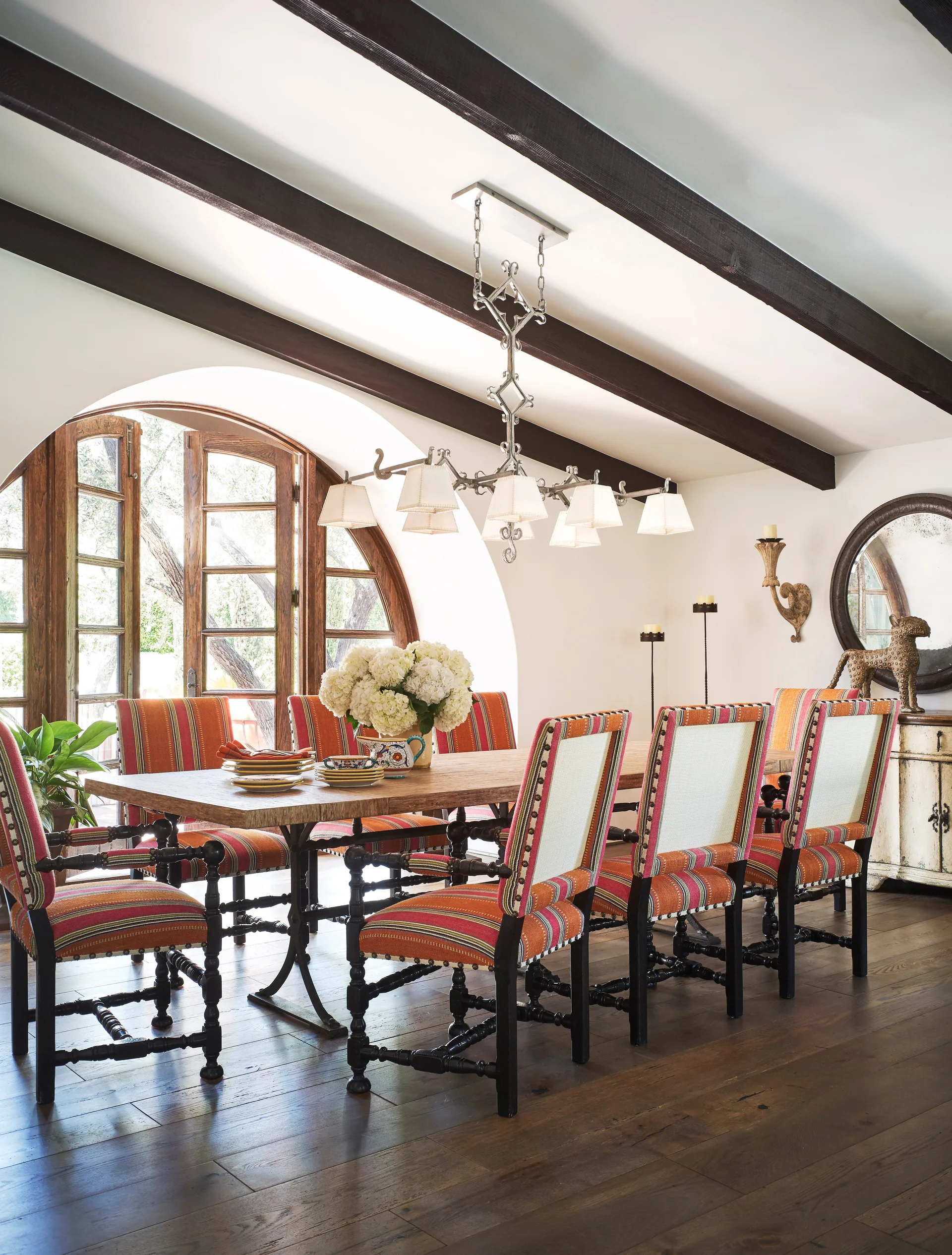 A rustic dining room features a long wooden table surrounded by striped upholstered chairs, dark ceiling beams, and a large arched window.