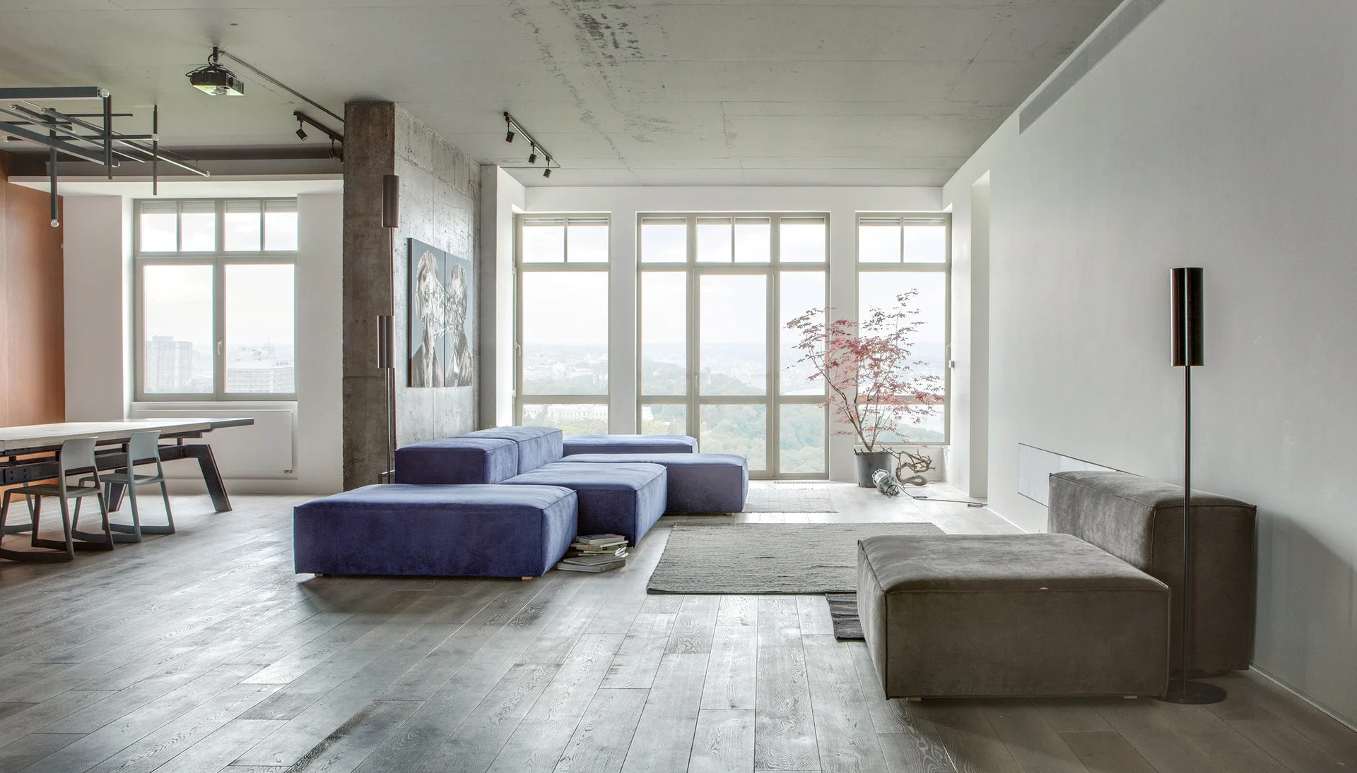 A minimalist open-plan living room features a low-profile blue modular sofa, an exposed concrete ceiling, and large windows.