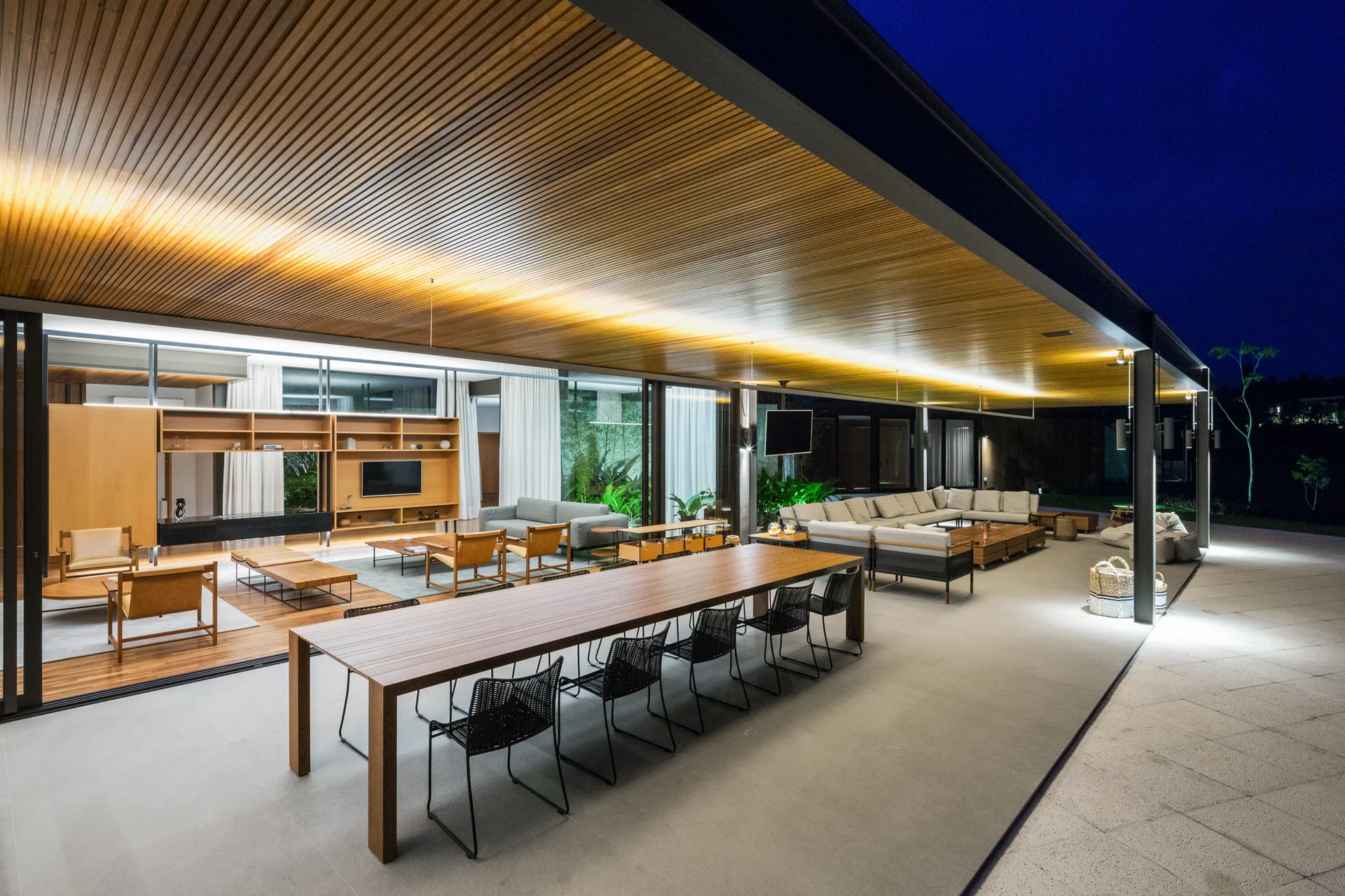 A spacious covered patio features a slatted wood ceiling with cove lighting, a long dining table, and a sectional sofa next to sliding glass doors.