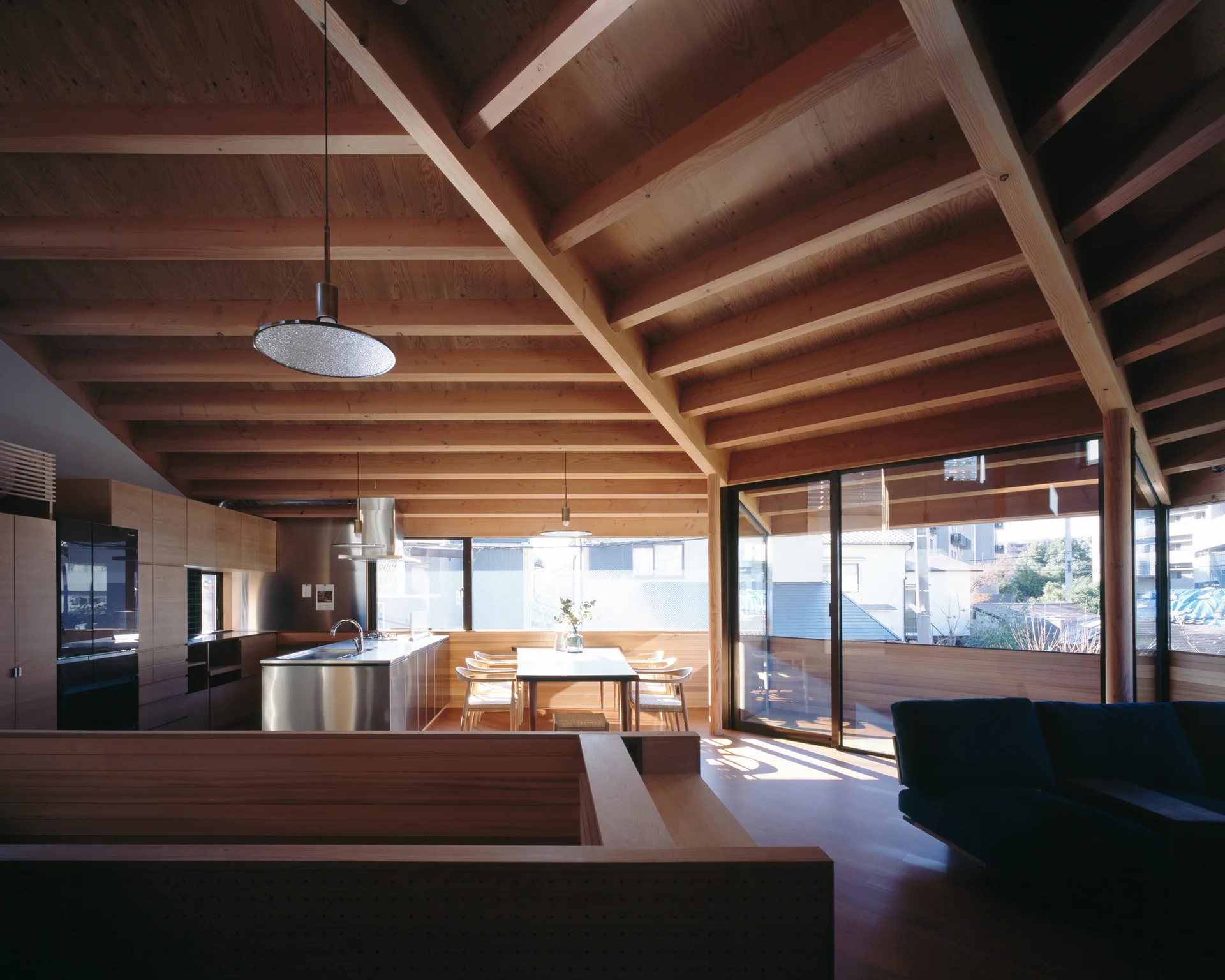 A modern open-plan home featuring a massive exposed timber ceiling, a sleek stainless steel kitchen island, and a dining table next to large glass doors.