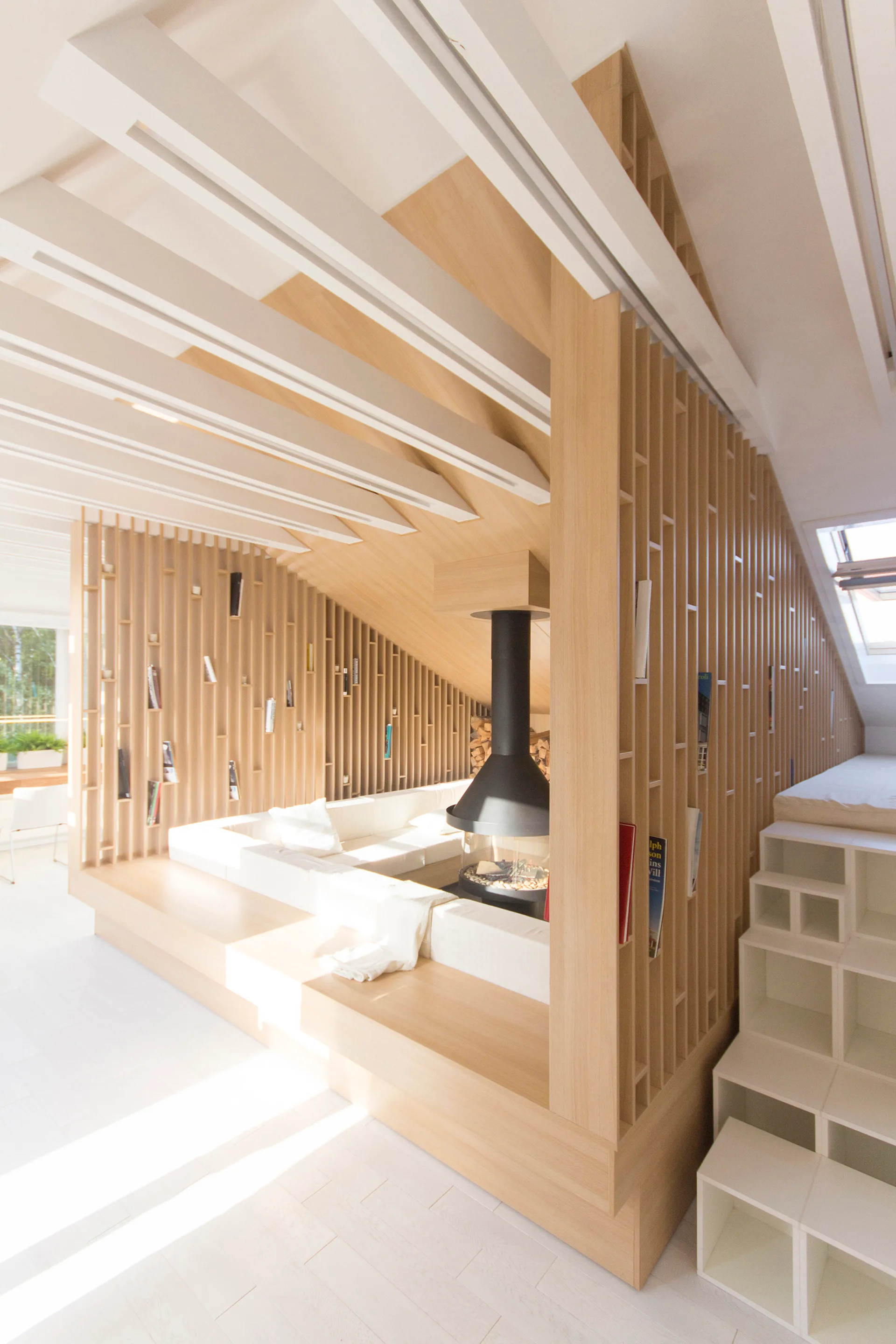 An attic living room featuring a white built-in sectional sofa, a suspended black fireplace, and a light wood slat partition wall.