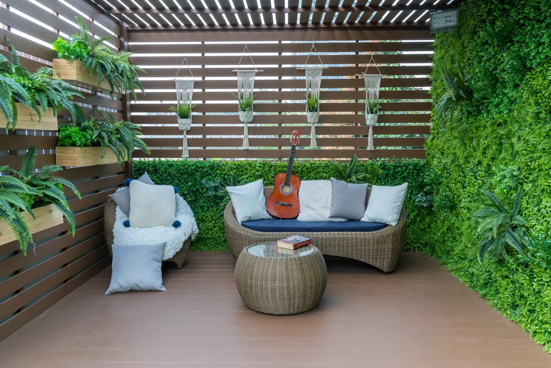 A covered patio with a wicker sofa and coffee table next to a lush green wall and horizontal wood slatted screen adorned with macrame plant hangers.