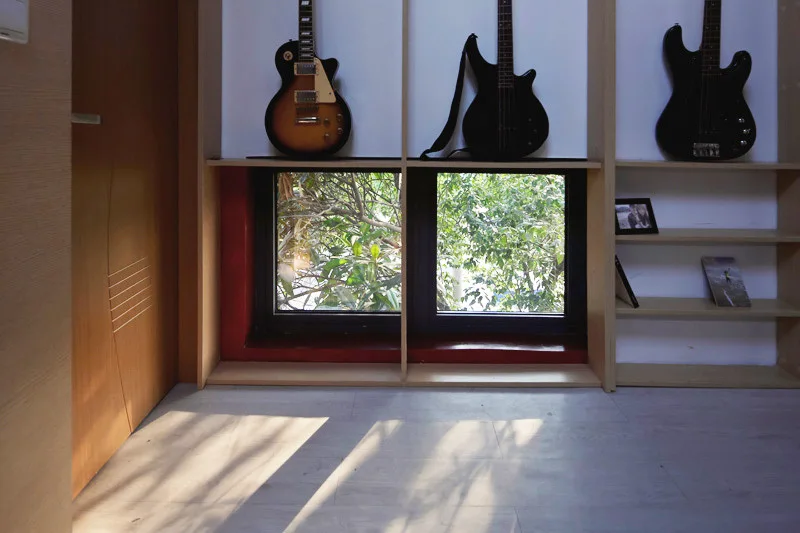 Light wood built-in shelving surrounds a low window showing green foliage outside, with three electric guitars displayed on the upper shelves.