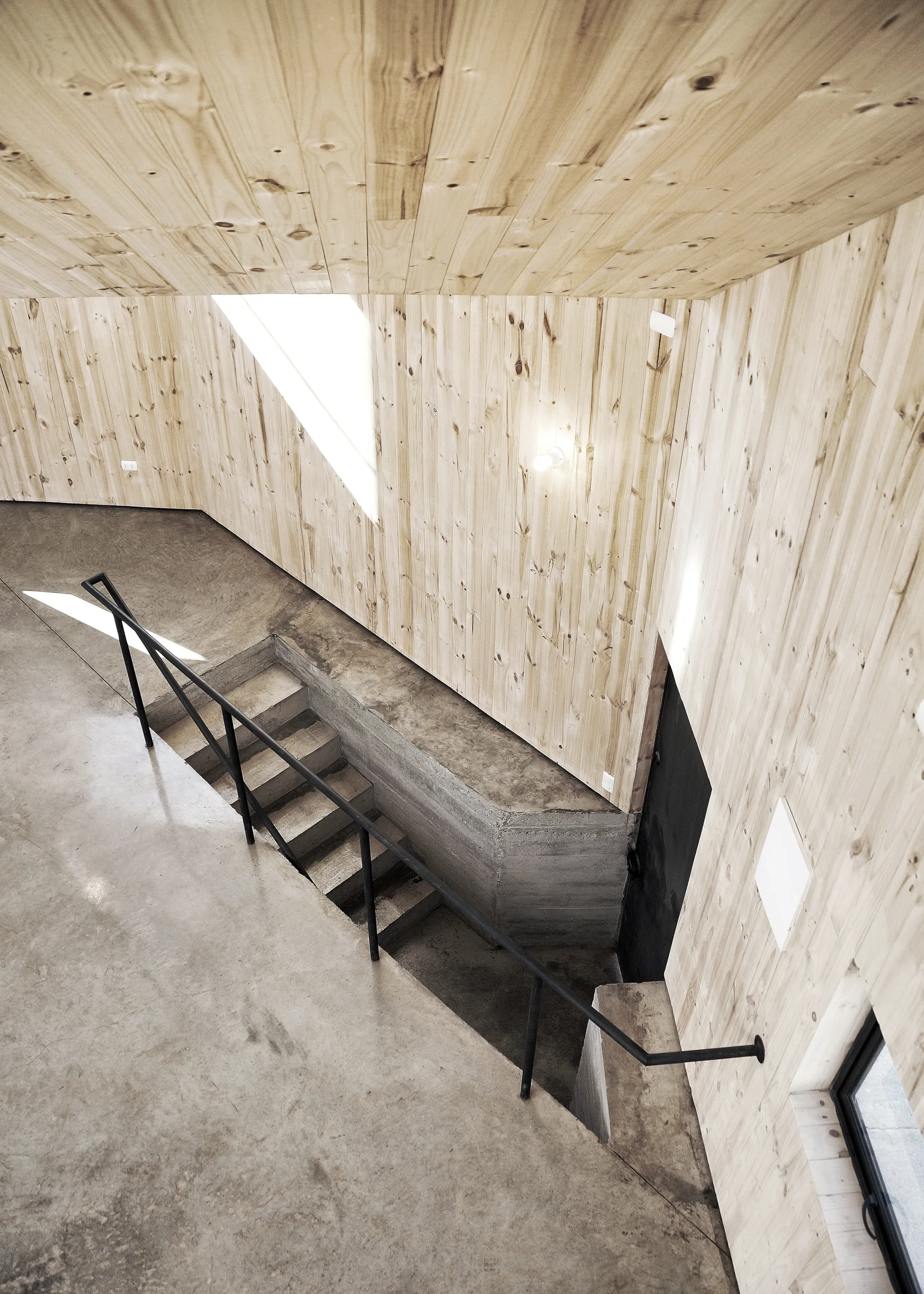 A concrete staircase with a black metal handrail is framed by light vertical wood paneling on the walls and ceiling.