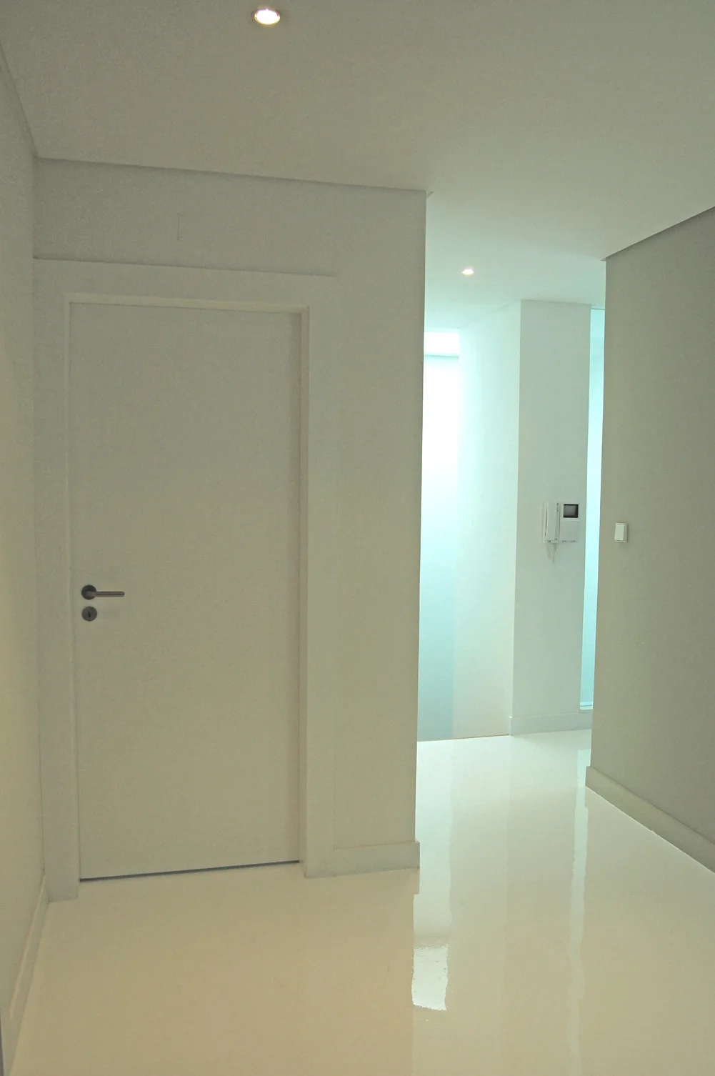 A minimalist white hallway features a glossy reflective floor, a simple white door with a dark handle, and recessed ceiling lights.